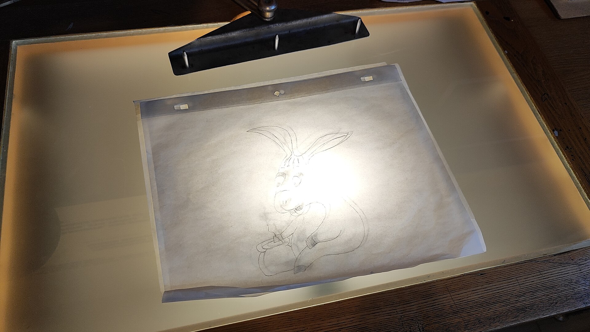A pencil sketch of a cartoon character on a translucent sheet lies on a backlit animation table under an angled lamp.