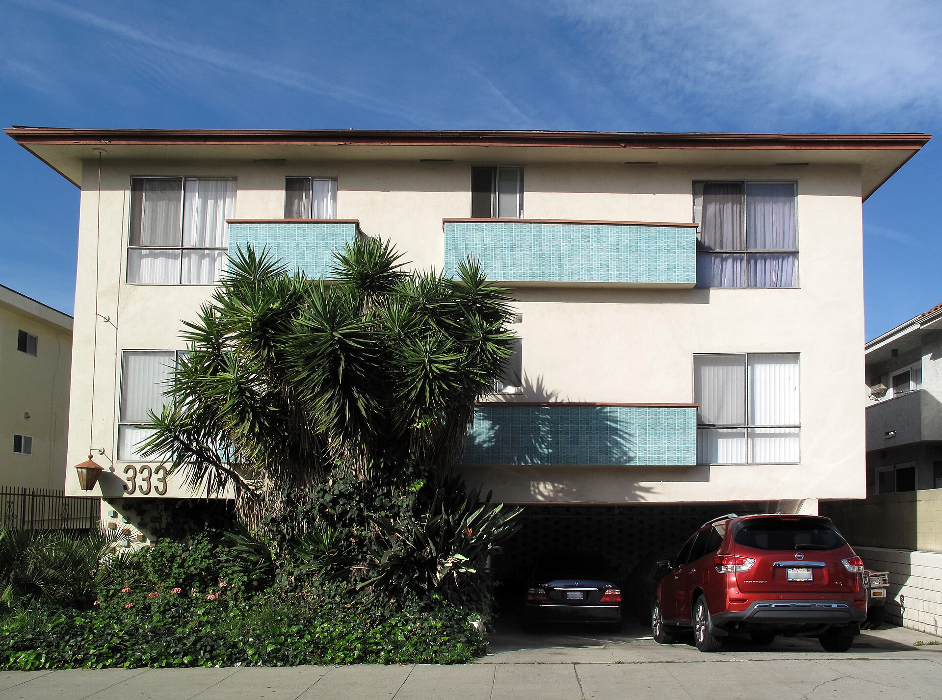 A three-story mid-century stucco apartment building with turquoise mosaic-tiled balcony panels and the number 333, a tall yucca in front and cars parked in the carport beneath, under a blue sky.