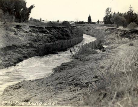 A swollen, muddy river running between eroded banks, a collapsed line of wooden fencing along the near side and houses among bare trees in the distance. Black-and-white archive photograph.