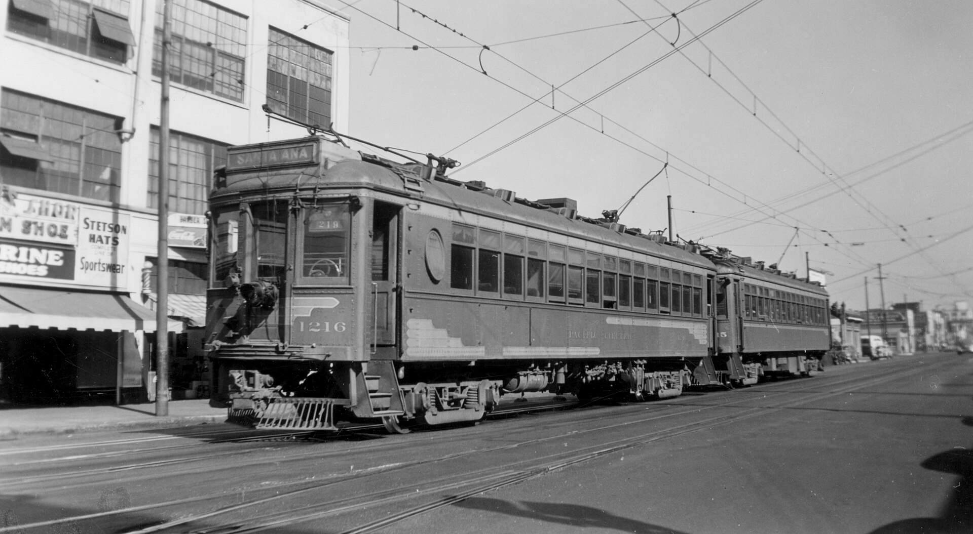 A two-car Pacific Electric interurban train, the front car numbered 1216 and signed for Santa Ana, running on street tracks past storefronts beneath a web of overhead wires. Black-and-white photograph, 1940s.