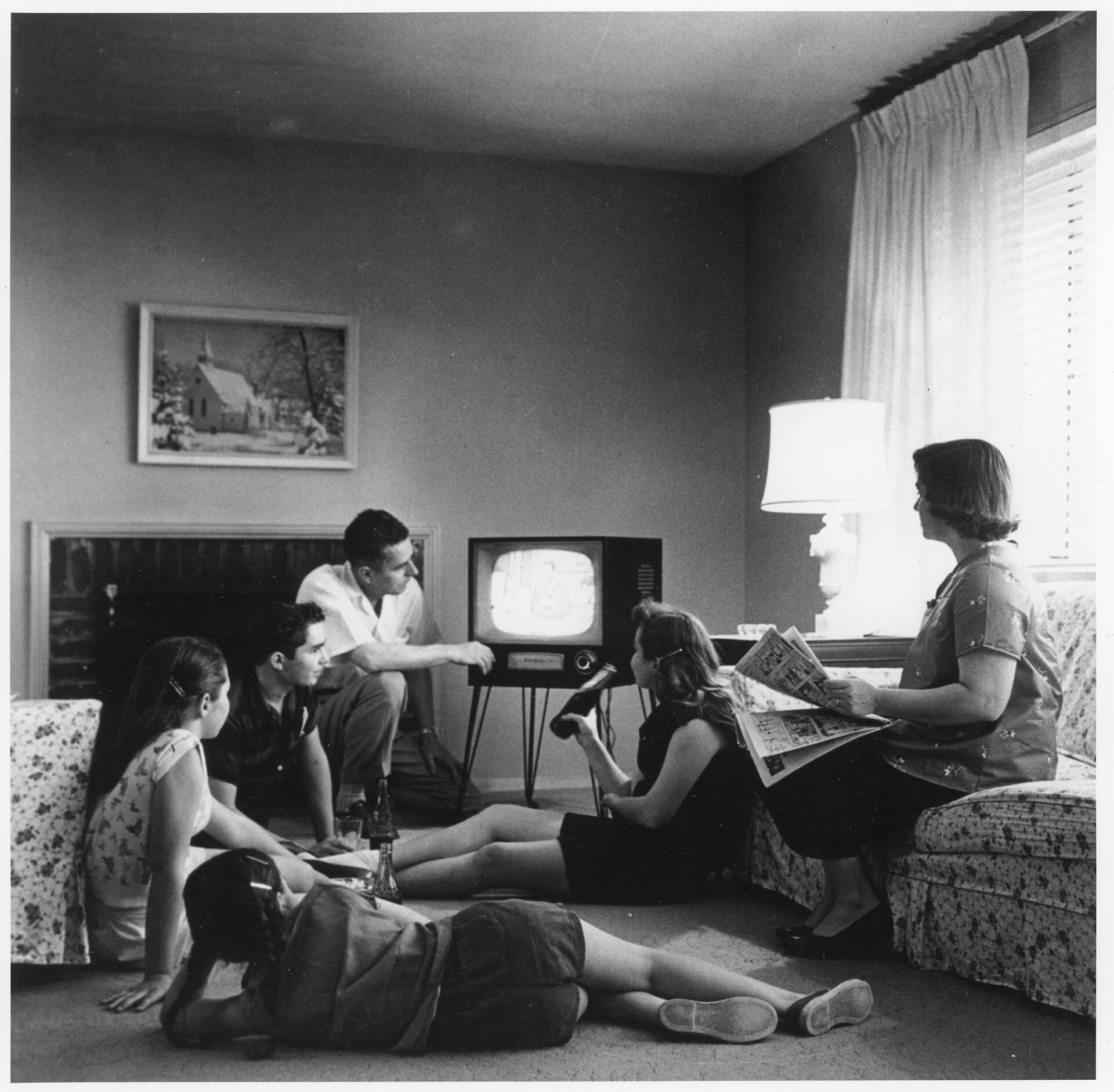 A family of six gathered in a living room around a small console television set, one girl reading a folded newspaper and soda bottles on the floor. Black-and-white photograph, 1950s.