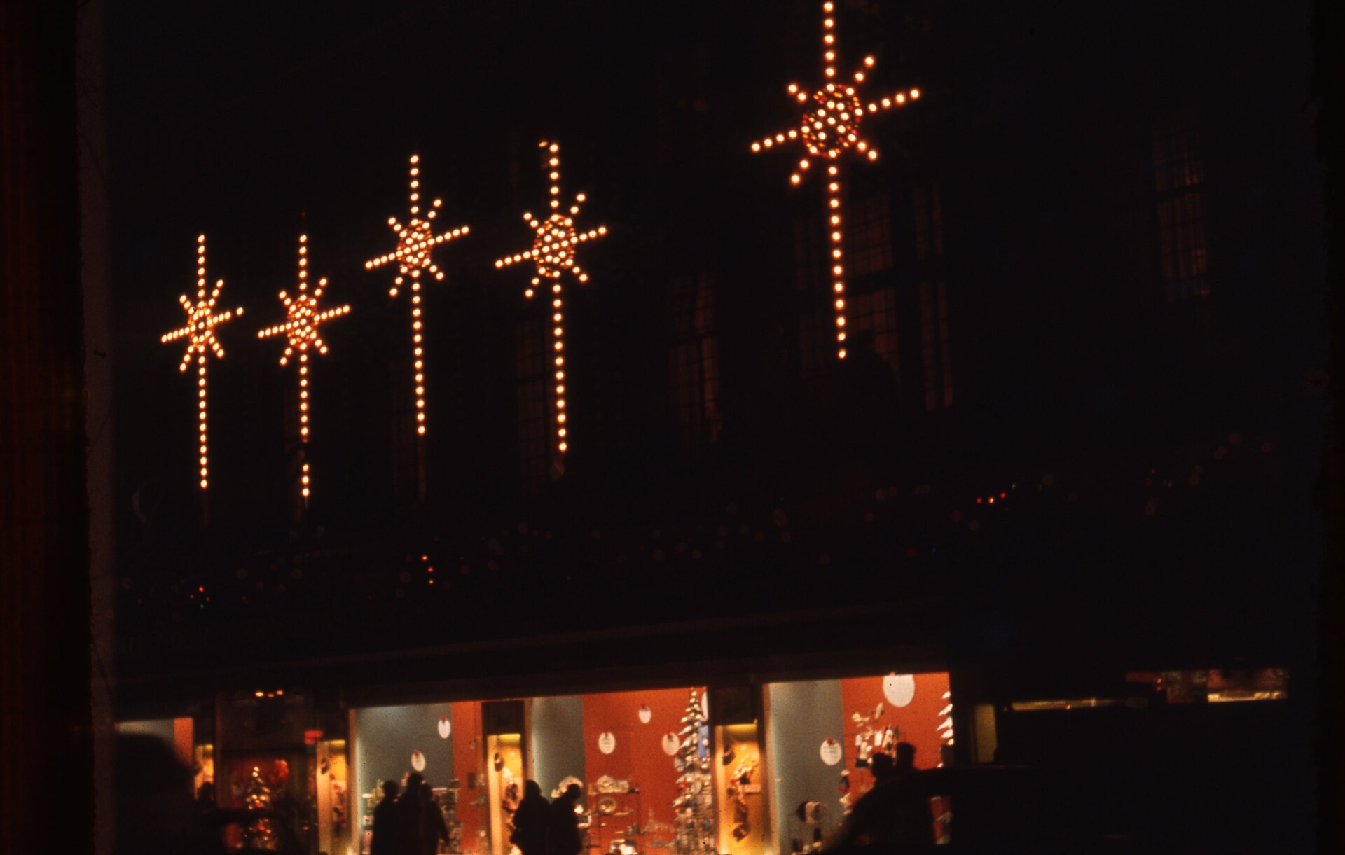 Illuminated star decorations glow above a lit department-store window at night, with Christmas displays inside and shoppers silhouetted on the dark sidewalk. Color photograph.