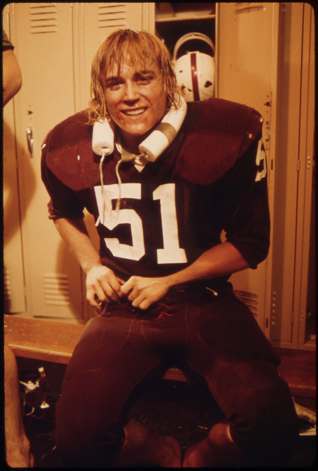 A young football player in a dark jersey numbered 51, hair damp and a roll of tape around his neck, sitting and smiling in front of a bank of metal lockers. Color photograph, 1970s.