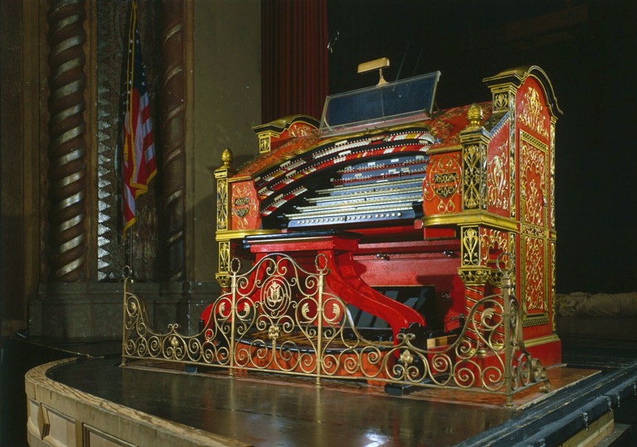 An ornate red-and-gold theatre pipe organ console with tiered keyboards and a curved brass railing, on a stage beside an American flag. Color photograph.