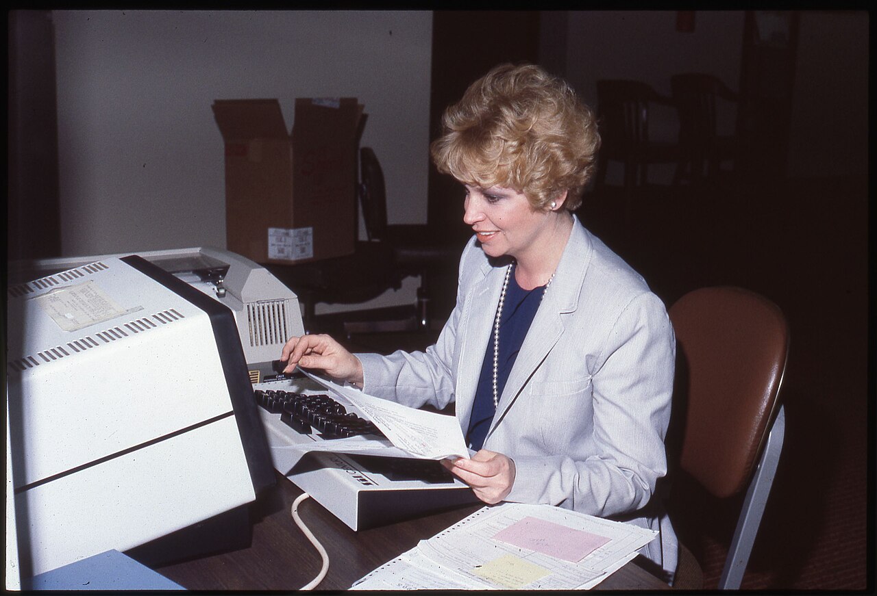 A woman in a pale blazer seated at a desk, one hand on the keyboard of a boxy desktop computer with a large monitor, holding a printed page. Color photograph.