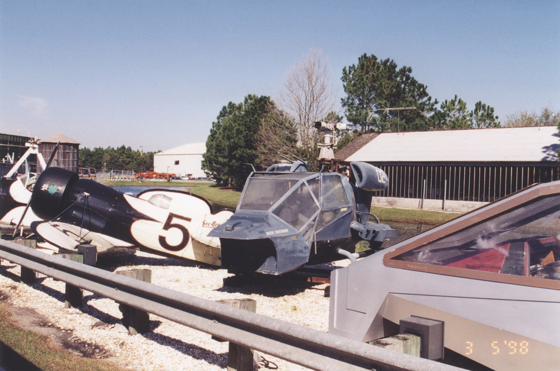 Retired picture vehicles sit outdoors on gravel at a studio backlot, including a black-and-white racing plane numbered 5 and a military-style helicopter mockup, with warehouse buildings and pines behind under a blue sky.