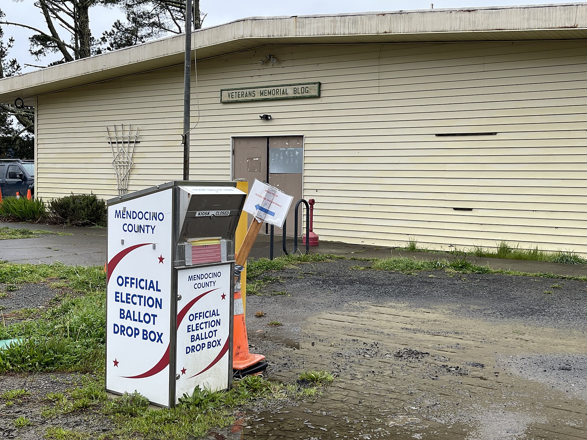 An outdoor official election ballot drop box stands on a grassy verge outside a single-story Veterans Memorial building, an orange traffic cone at its base.
