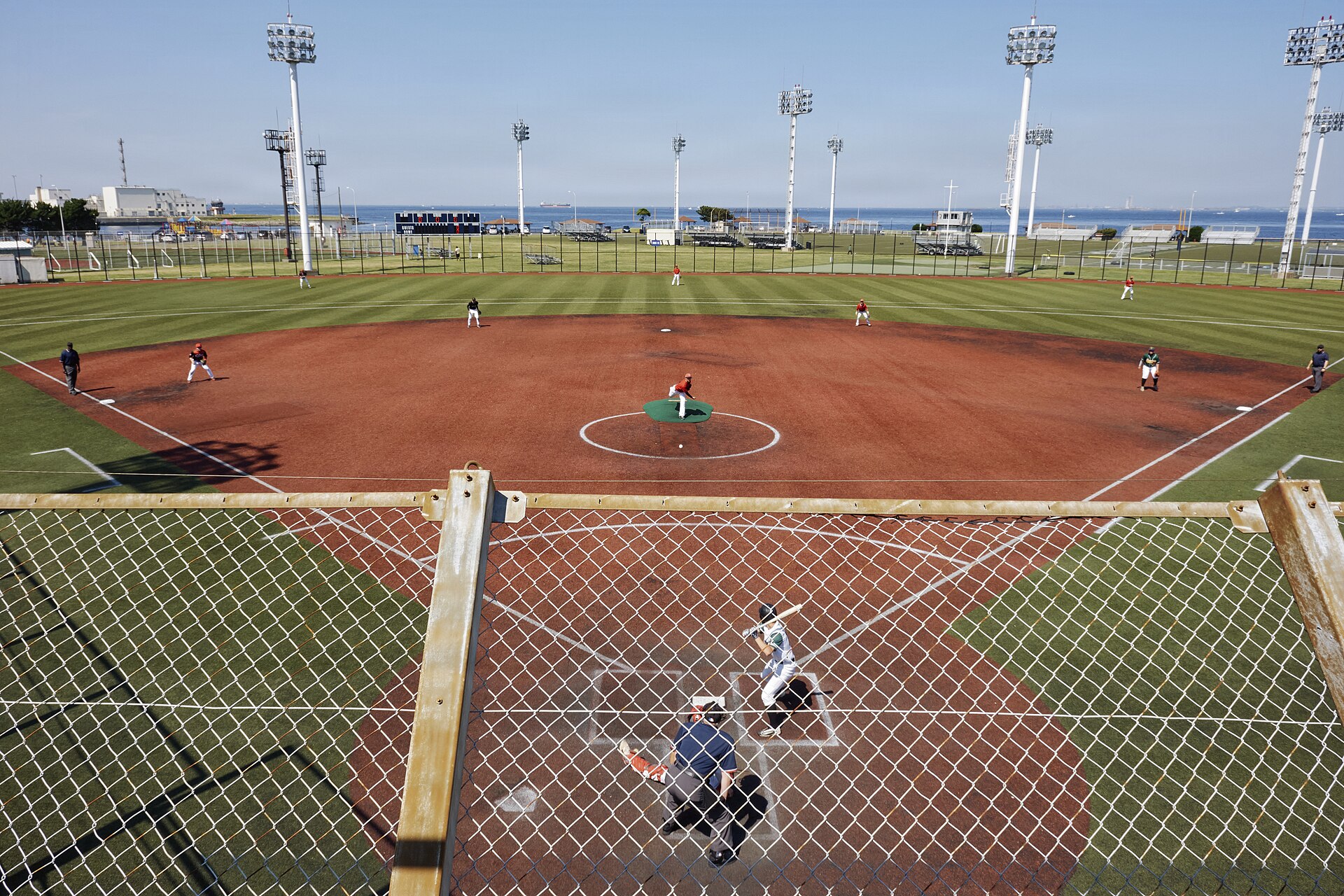 A batter, catcher and umpire at home plate seen through the backstop netting during a baseball game, with fielders positioned on the field and the sea beyond the outfield fence.