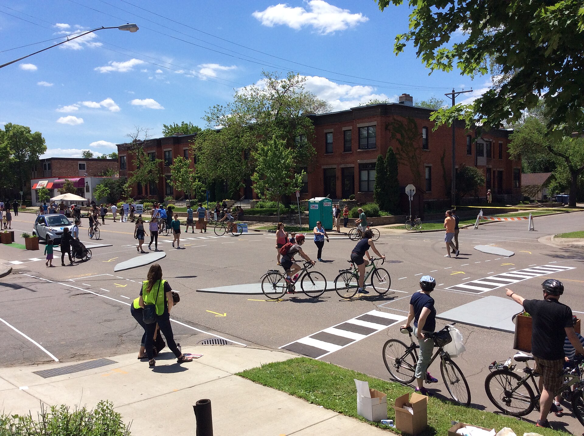 People walk and ride bicycles along a residential street closed to cars for an open-streets event, with temporary painted crosswalks and curb markings and brick rowhouses behind.