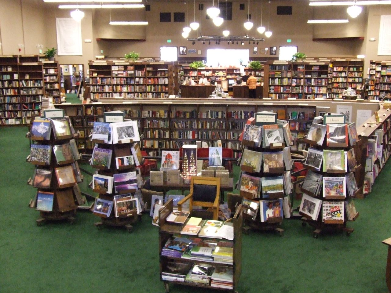 The interior of a spacious independent bookstore with green carpet, rotating racks of books and calendars, and full floor-to-ceiling shelves along the walls.