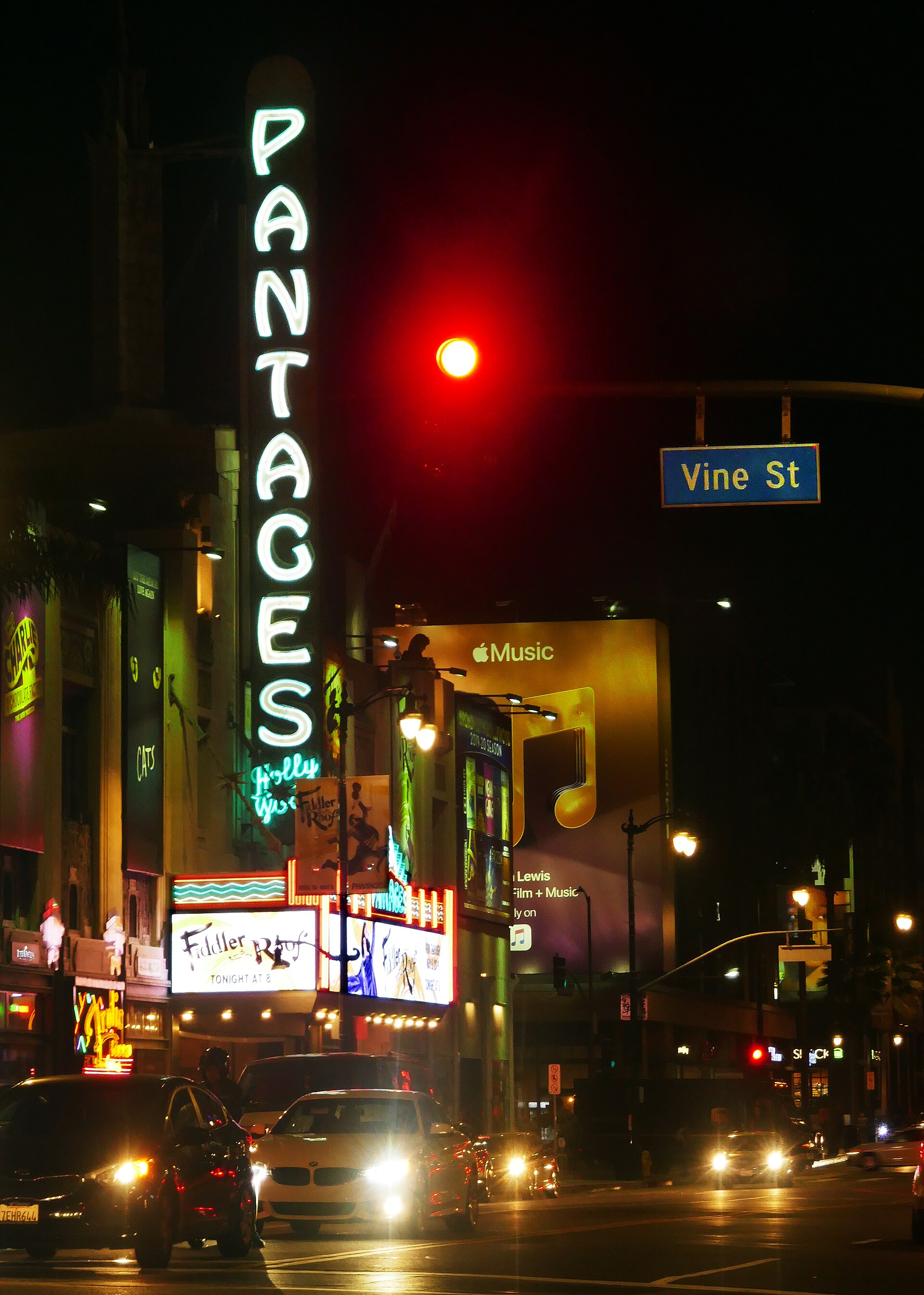 The illuminated vertical PANTAGES sign and theater marquee on Hollywood Boulevard at night, with a Vine Street sign, a red traffic signal and headlights of passing cars.