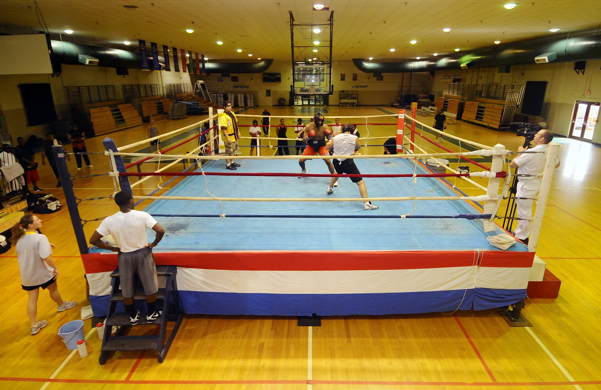 Two boxers in protective headgear spar inside a roped ring on a gymnasium floor as onlookers and a cameraman watch from the sides.
