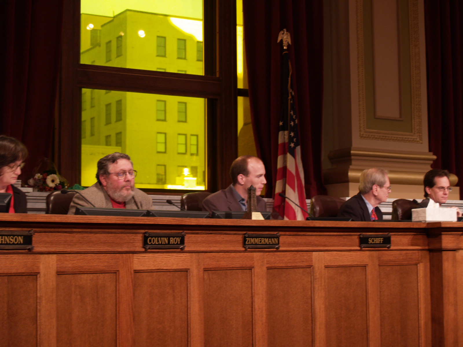 Five council members sit at a long wooden dais with engraved brass nameplates during a meeting, a U.S. flag and heavy red curtains behind them and a lit building visible through a tall window.