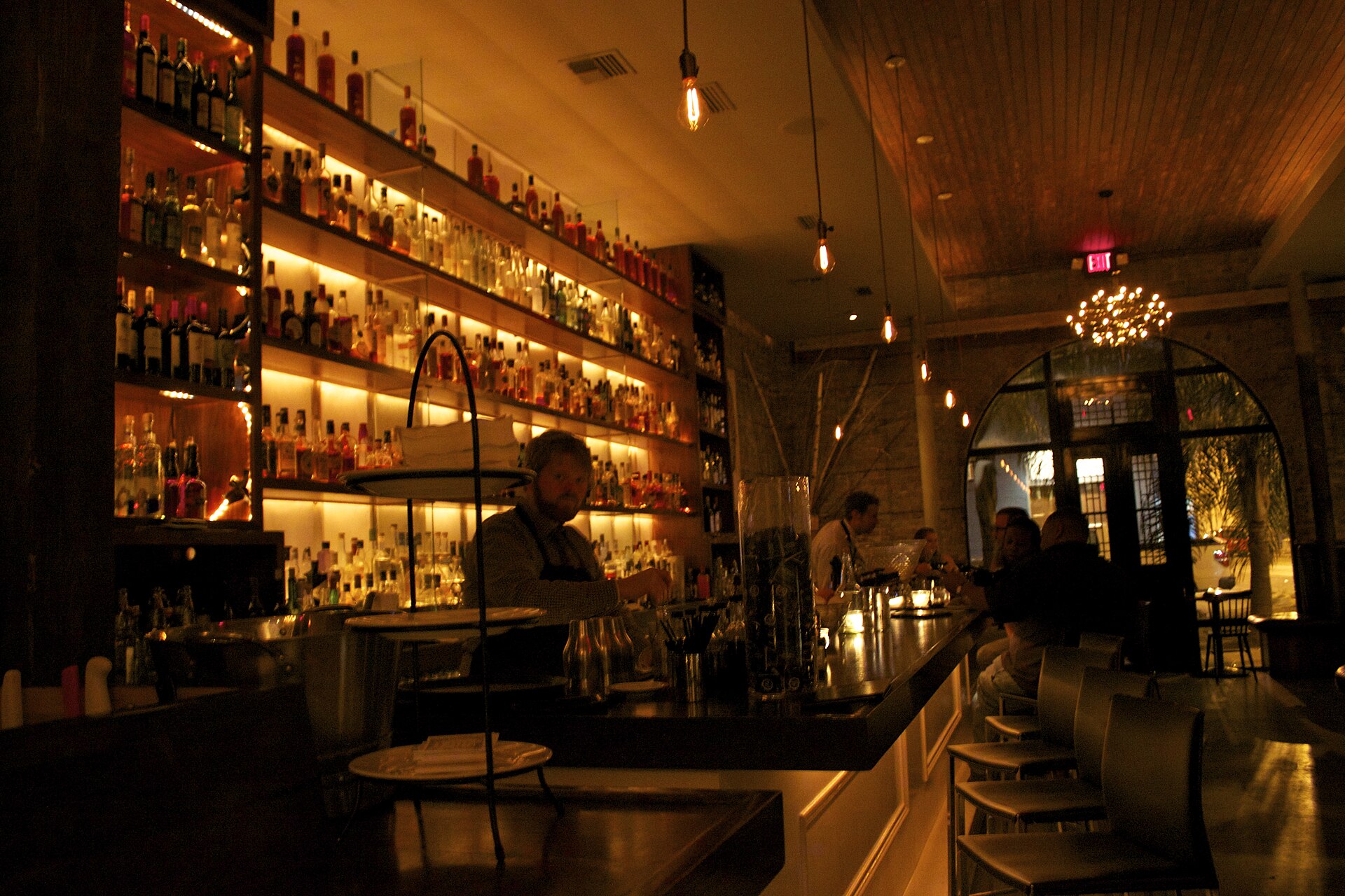 A bartender in an apron works behind a long, dimly lit bar backed by warmly lit shelves stacked with liquor bottles, bare-filament bulbs hanging overhead.