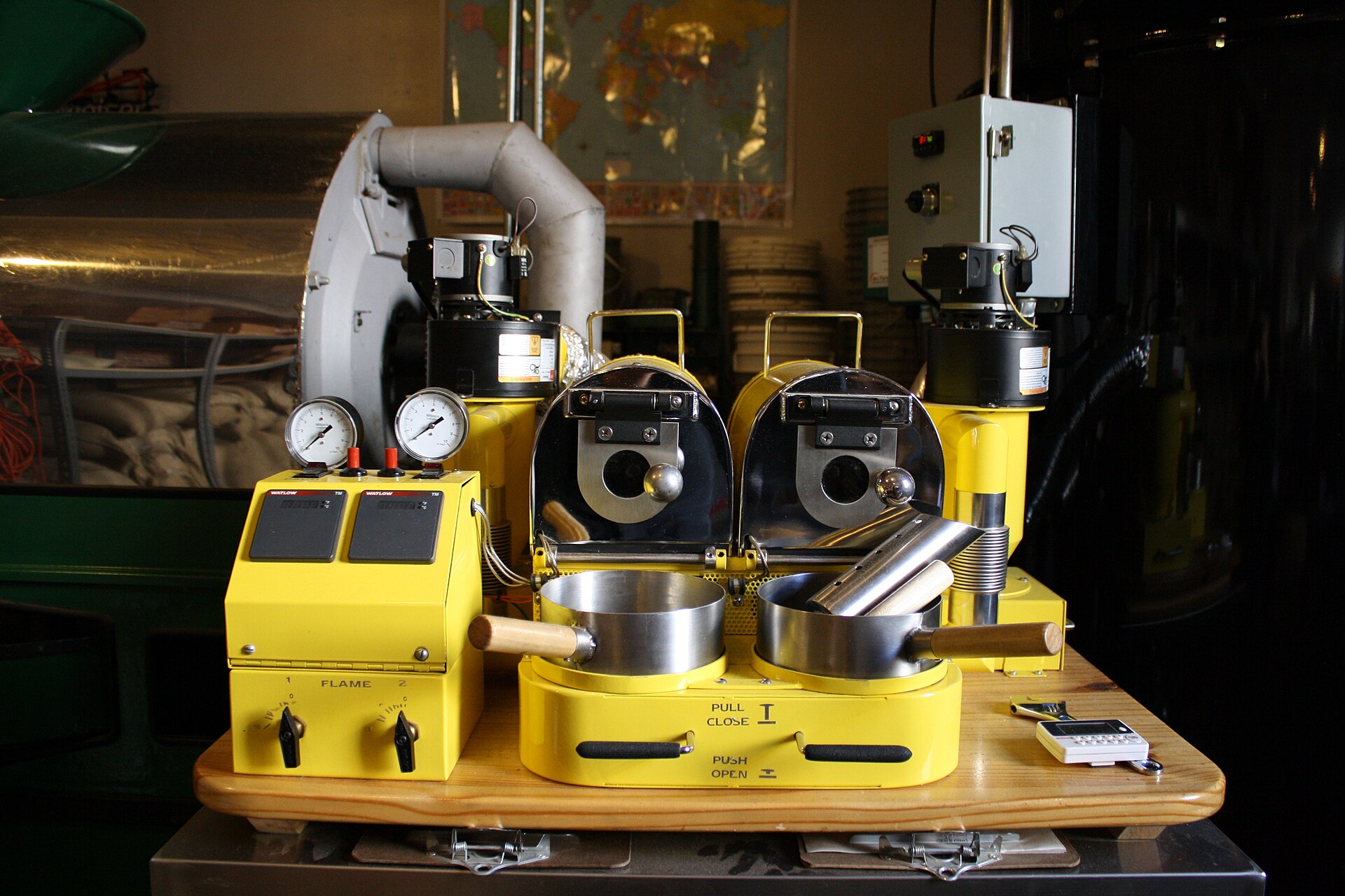 A bright yellow twin-drum coffee roasting machine with temperature gauges and cooling trays, burlap sacks of green coffee beans and a world map behind it.