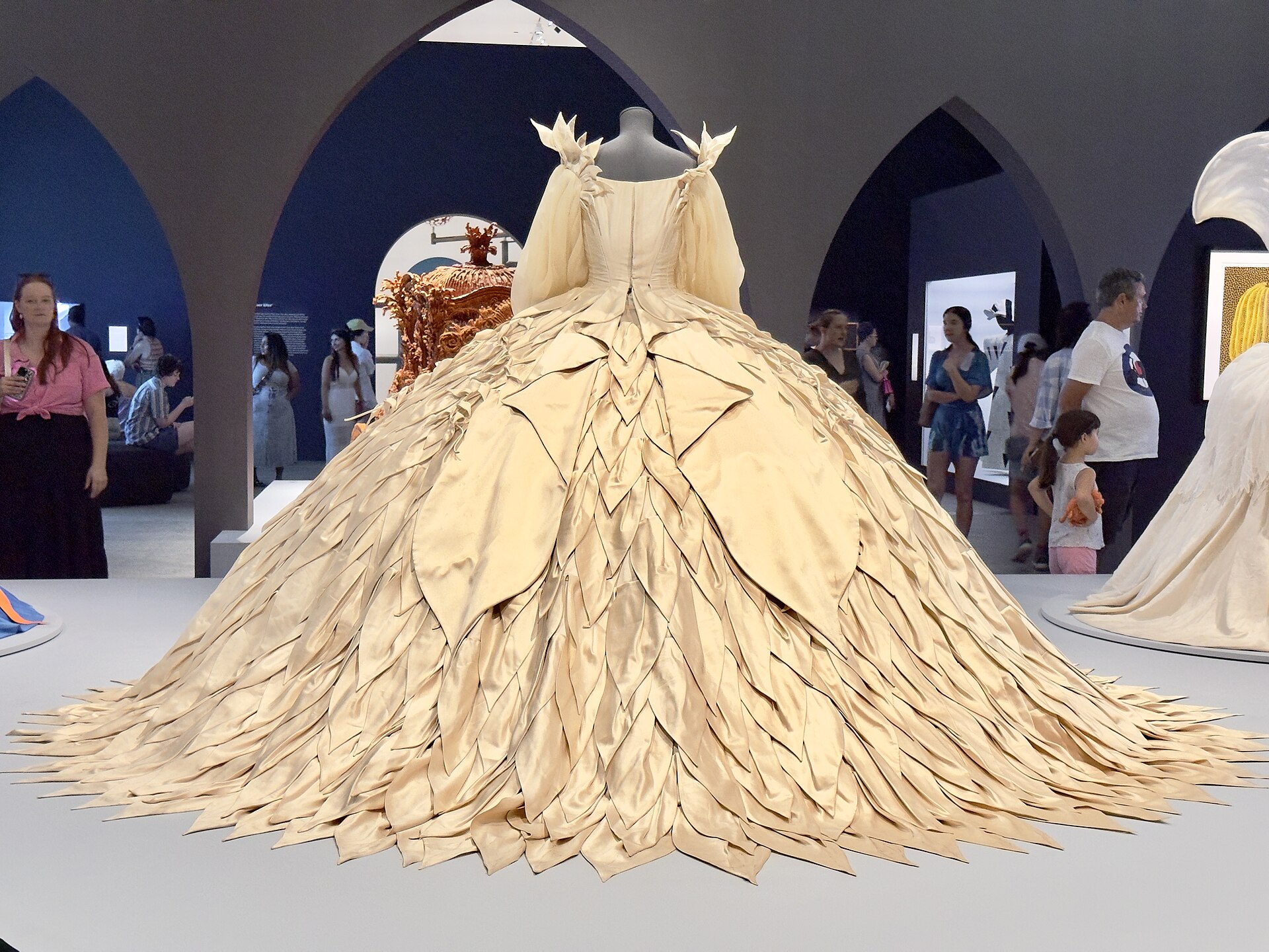 A voluminous cream satin ball gown with a layered, petal-like skirt on a mannequin, displayed on a low platform in a museum gallery as visitors pass under dark pointed archways.