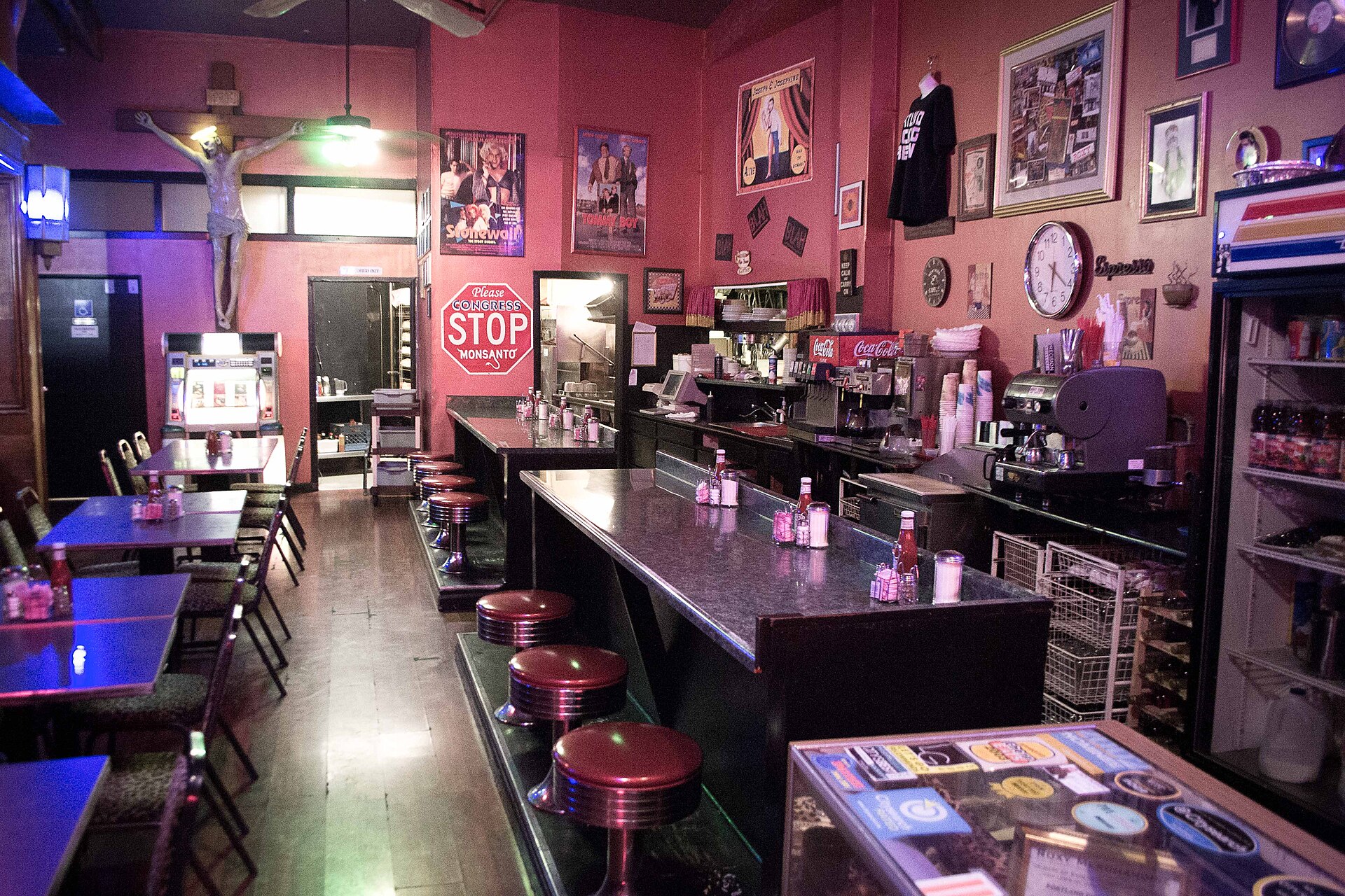 The counter and empty red-topped stools of a dimly lit vintage diner, its walls crowded with framed posters, a clock and a soda fountain.