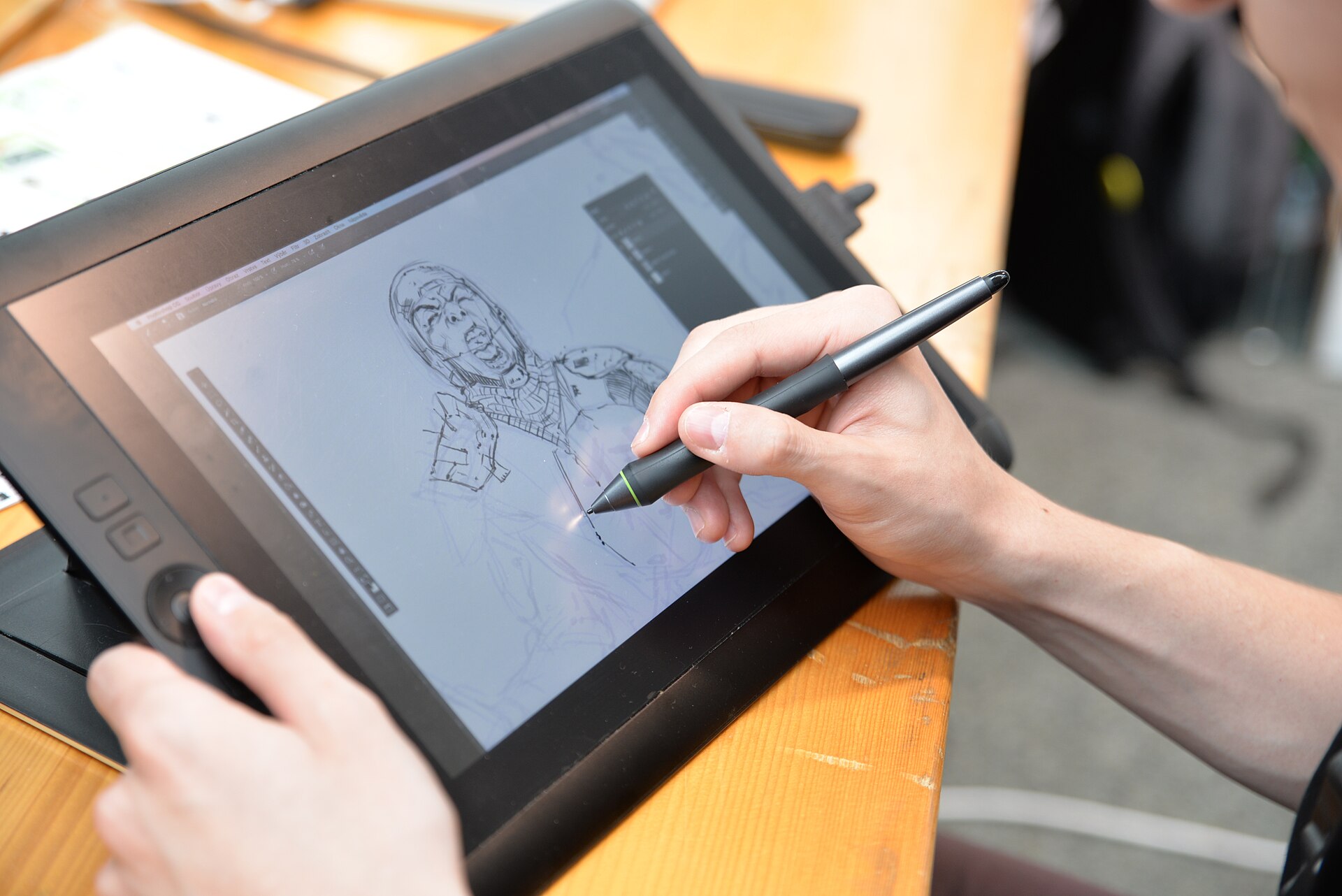 A pair of hands holds a stylus to a pen-display tablet on a wooden desk, sketching a helmeted character in a drawing program on the screen.