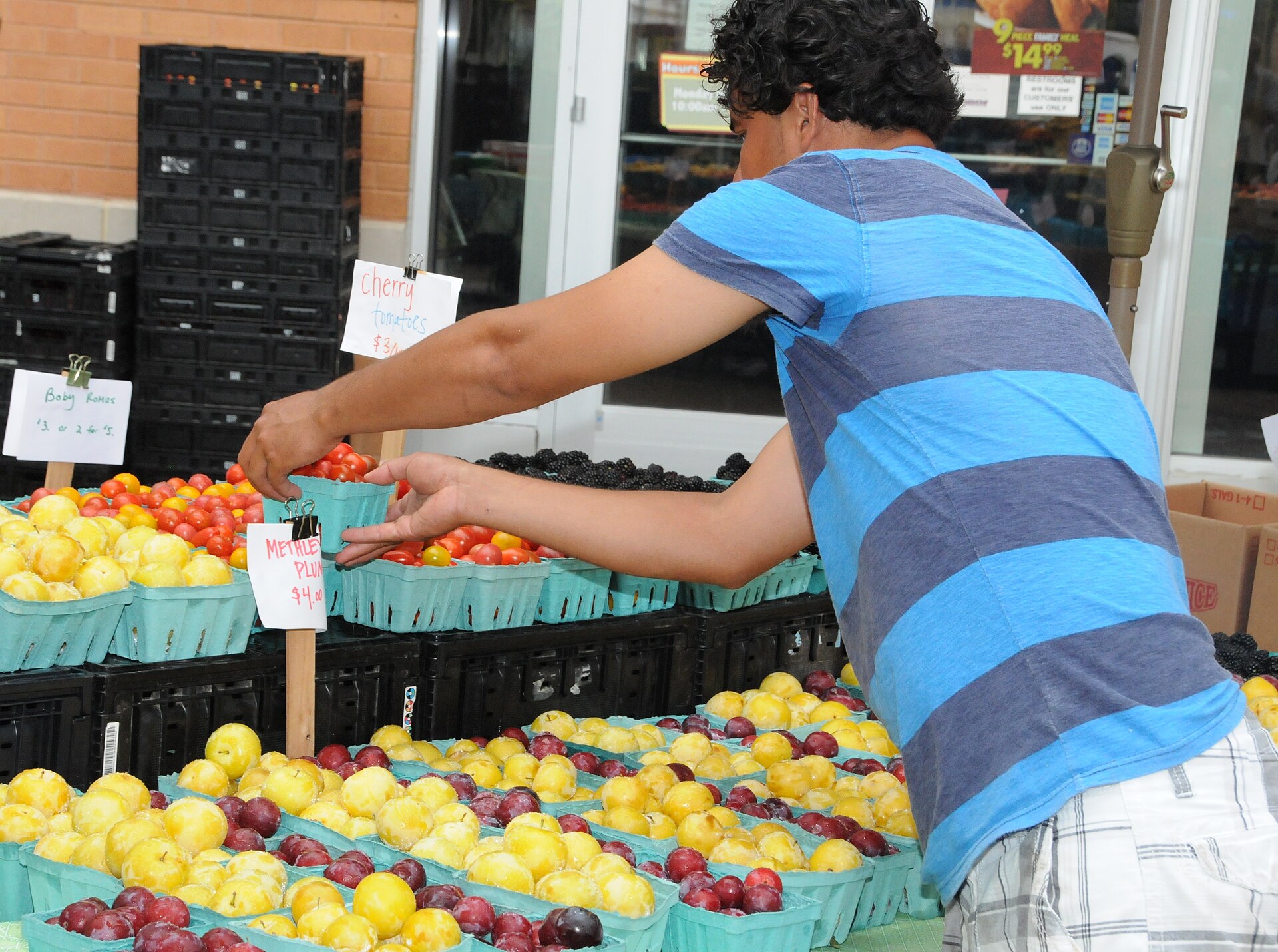 A person in a striped shirt reaches across a produce stand piled with baskets of yellow and red plums and cherry tomatoes, under hand-lettered price signs.