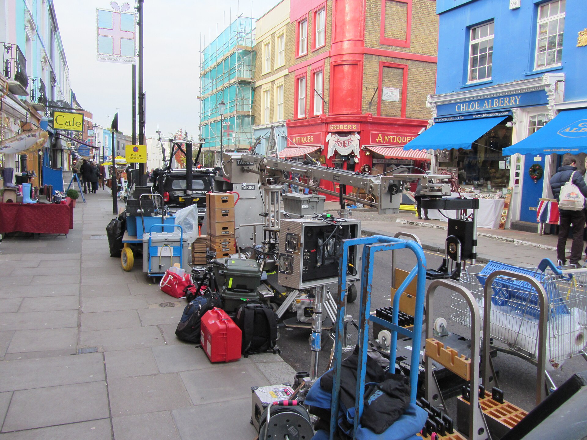 Grip carts, road cases and a camera crane crowd the sidewalk of a colorful shopping street during a location film shoot.