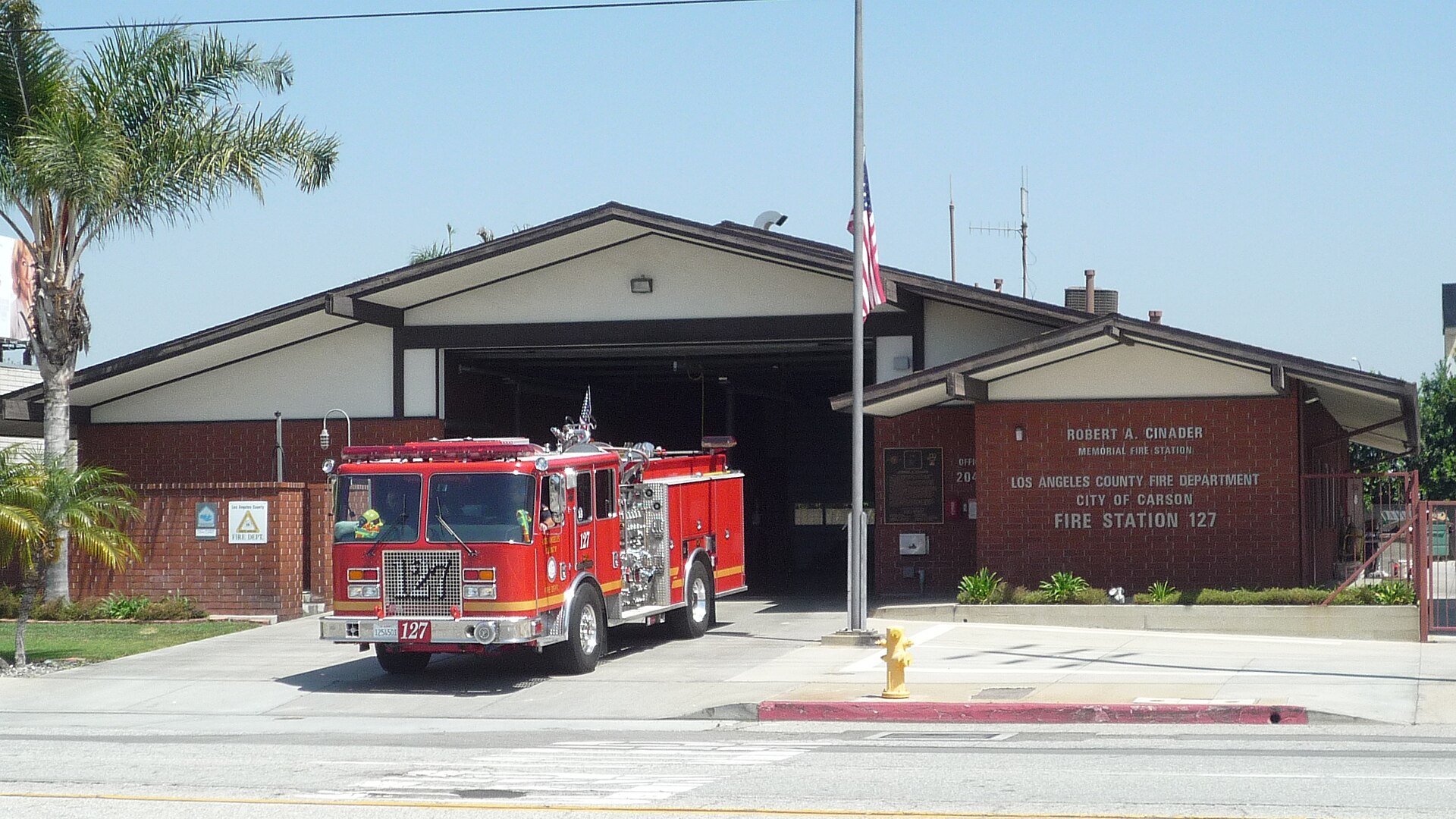 A red fire engine parked in the driveway of a single-story brick fire station on a sunny day, with a palm tree and a flag on a pole beside the open apparatus bay.