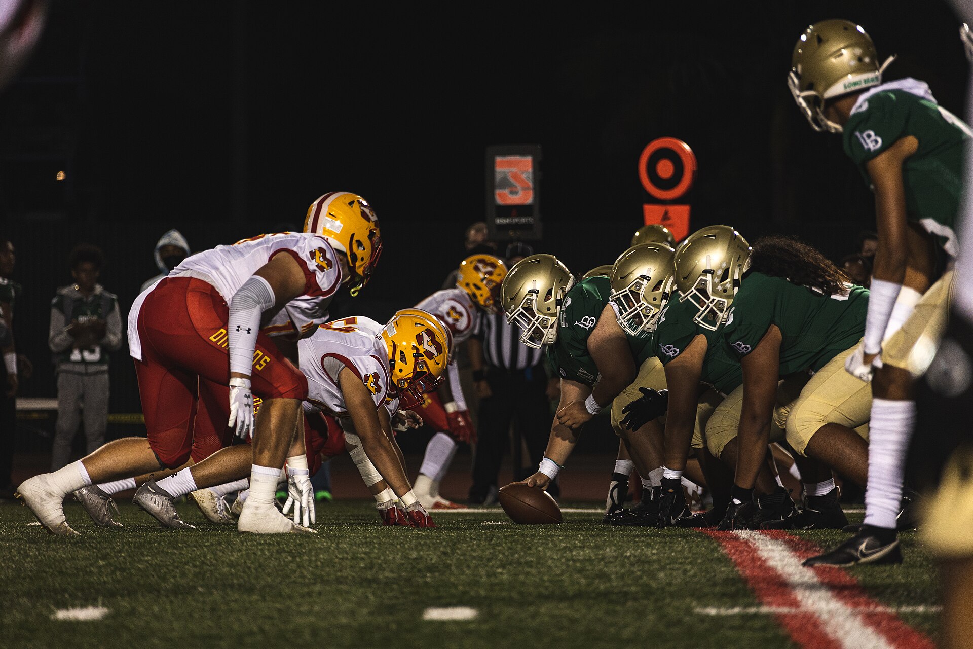 Two lines of helmeted high-school players crouch face to face at the line of scrimmage under floodlights at night, the center's hand resting on the ball, with a striped official and spectators dimly visible behind them.