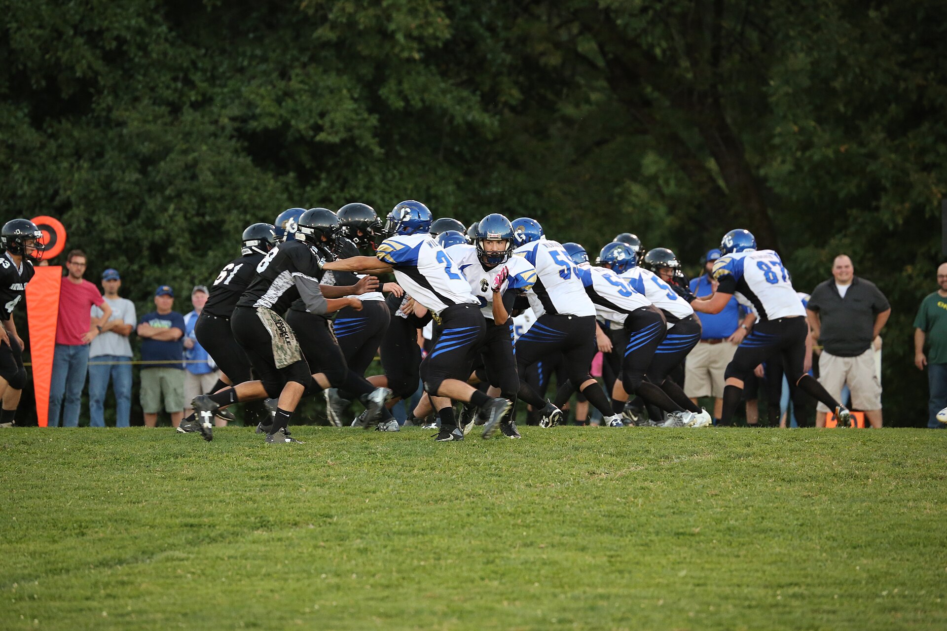 Two high school football teams in helmets and pads meet at the line of scrimmage on a grass field, spectators watching from behind a sideline rope.