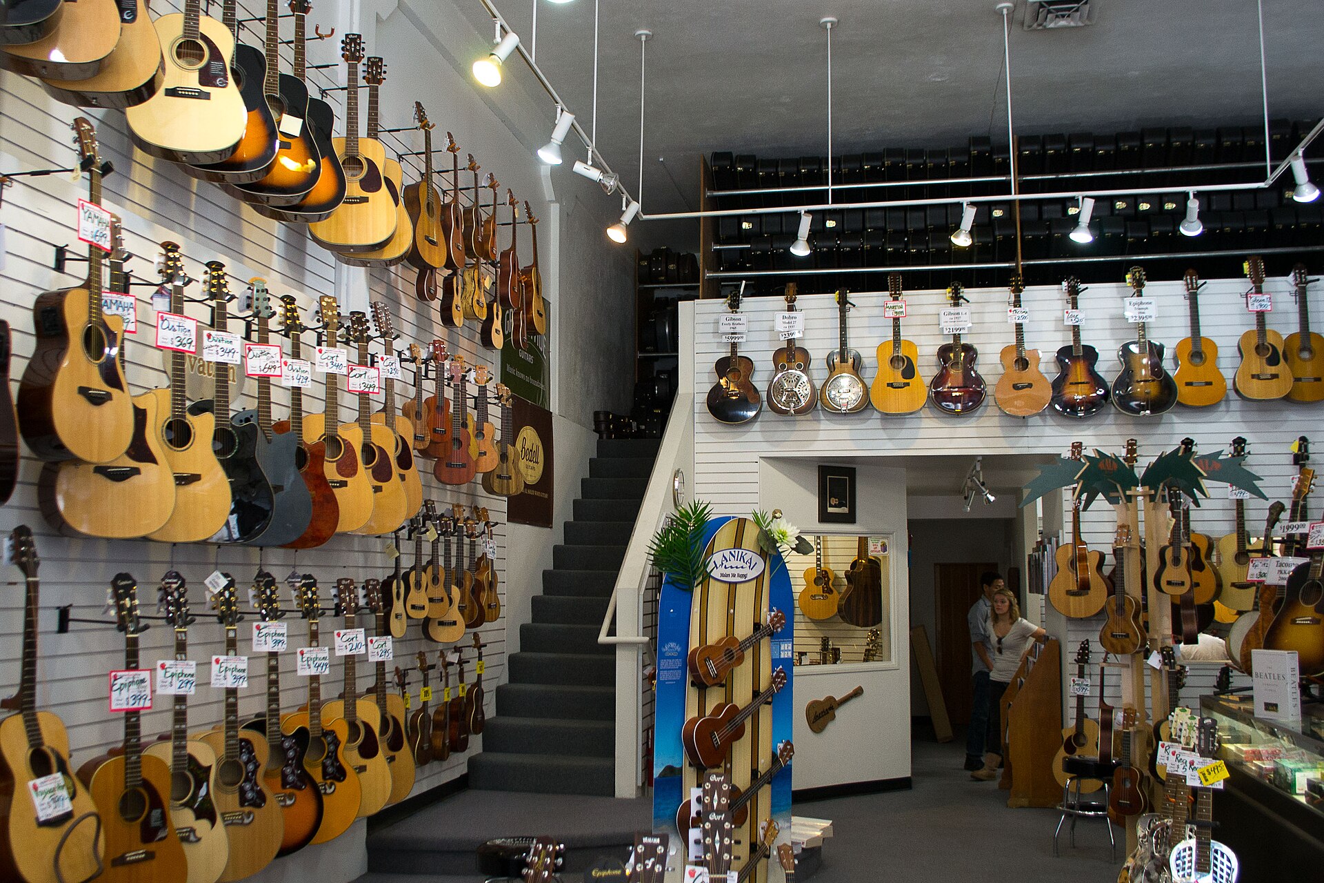 The walls of a guitar shop densely hung with acoustic and electric guitars and ukuleles above a carpeted staircase, price tags on each and a glass display counter to the right.