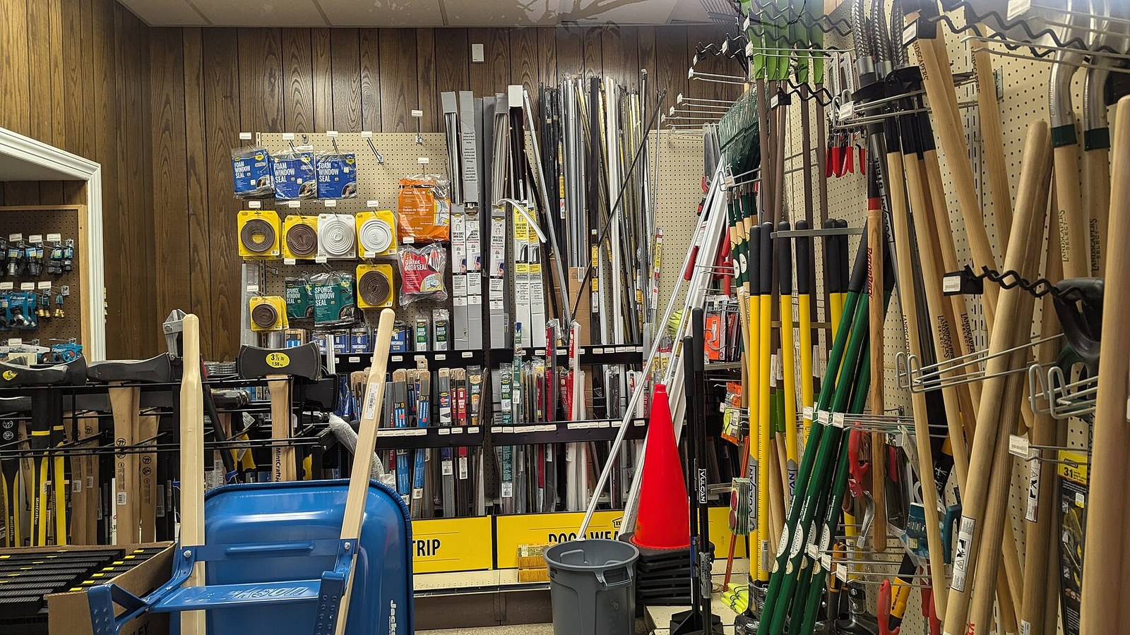 The inside of a small hardware store: wood-panelled walls and pegboard hung with weatherstripping packets, axes, rakes and long-handled garden tools, with a blue wheelbarrow, a grey trash can and an orange traffic cone in the foreground.