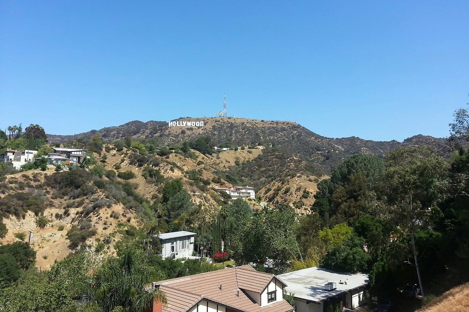 Houses sit among trees on dry, scrub-covered hillsides under a clear blue sky, with the Hollywood Sign on the ridge in the distance.
