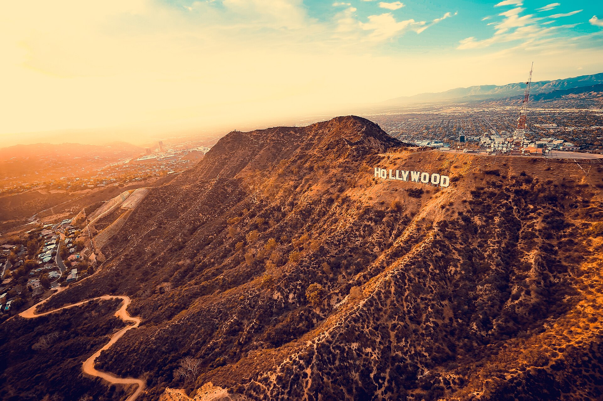 An aerial view at golden hour of the Hollywood Sign on the brown-and-green flank of Mount Lee, with a dirt trail winding up the hillside and the city spread out beyond.