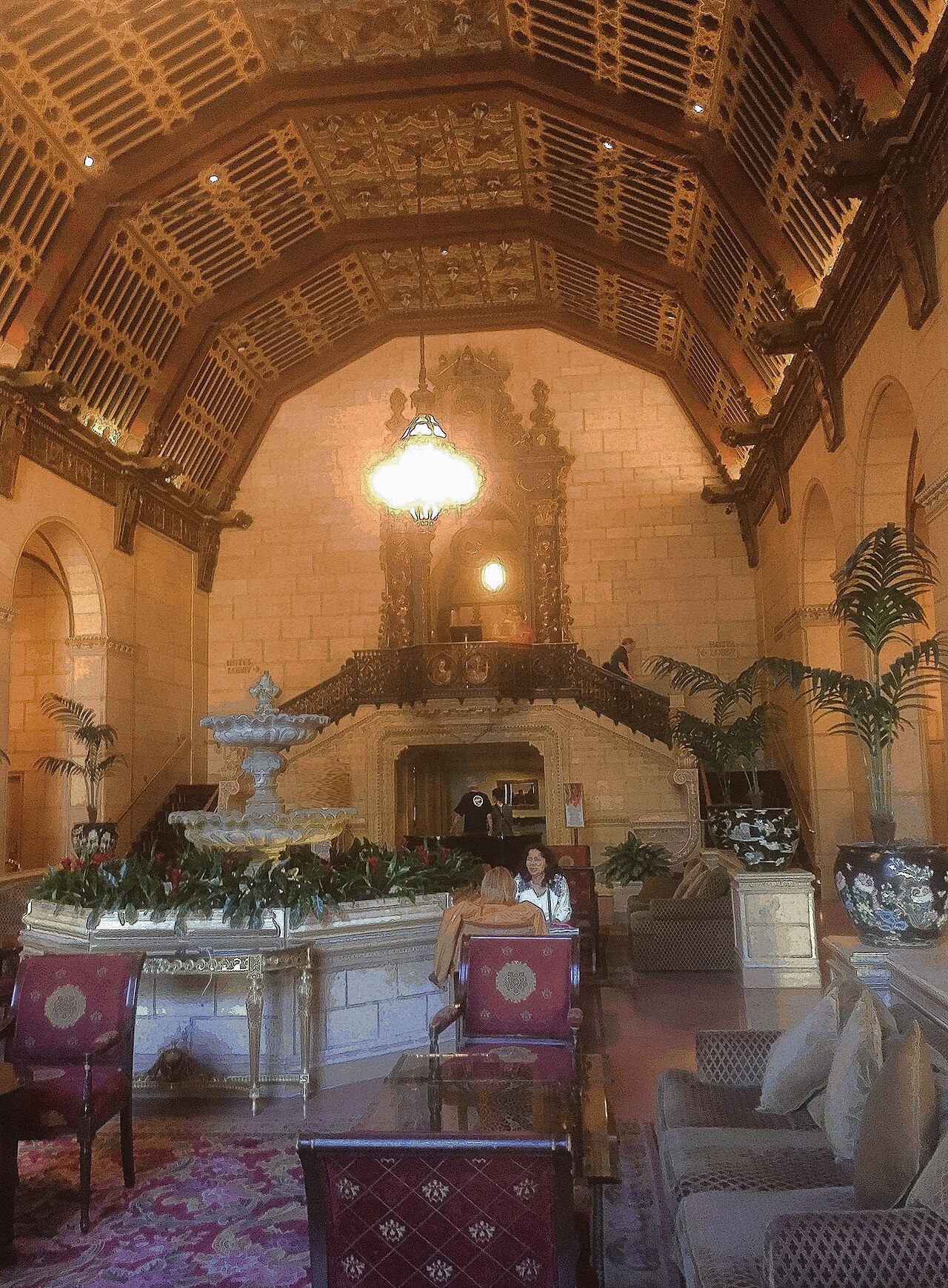 The ornate lobby of a historic hotel with an elaborately carved and painted vaulted ceiling, a large hanging lamp, a tiered stone fountain ringed with greenery, potted palms and patterned armchairs.