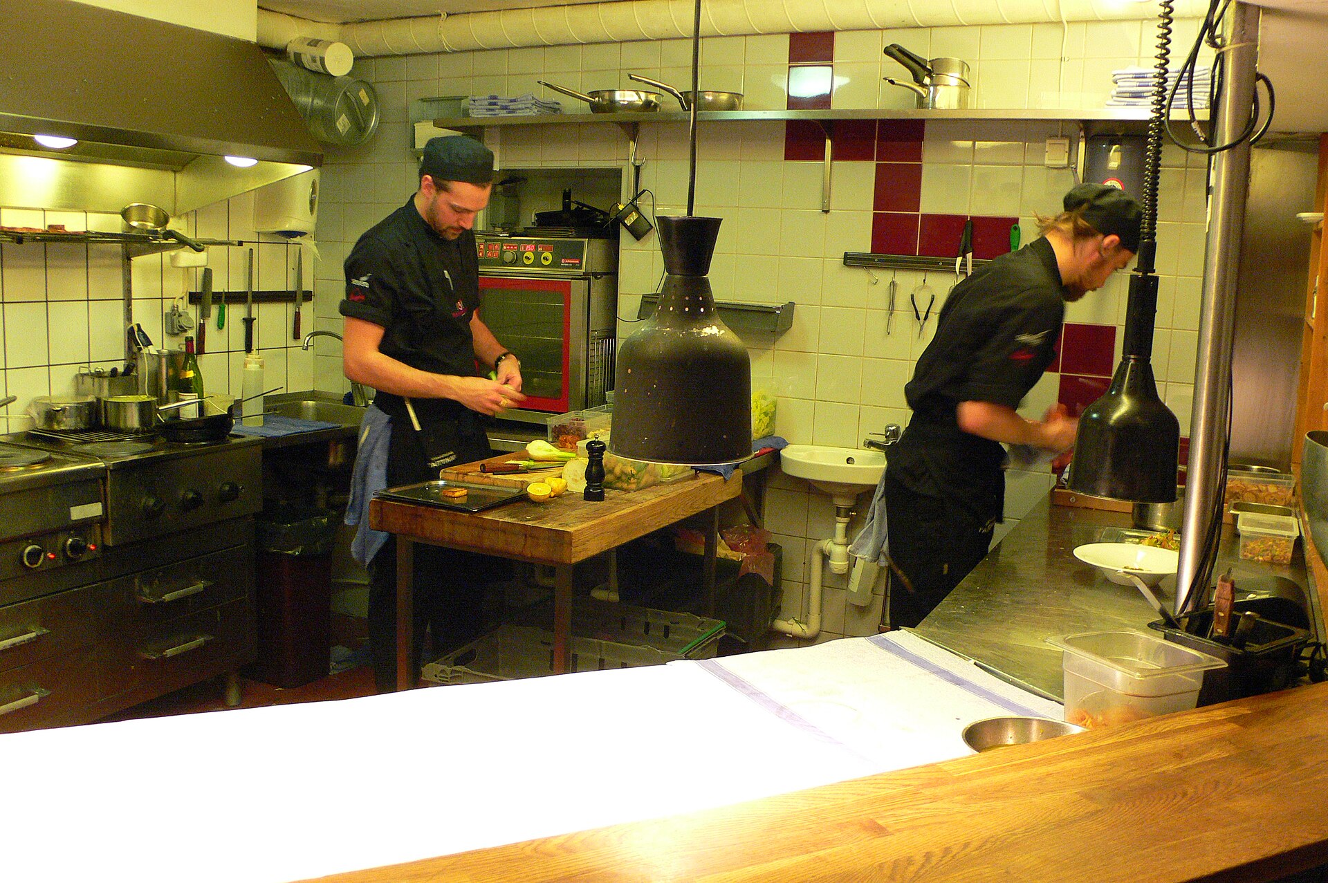 Two cooks in black chef's jackets and caps work in a small stainless-steel restaurant kitchen, one slicing at a wooden butcher block, the other plating under warming lamps.