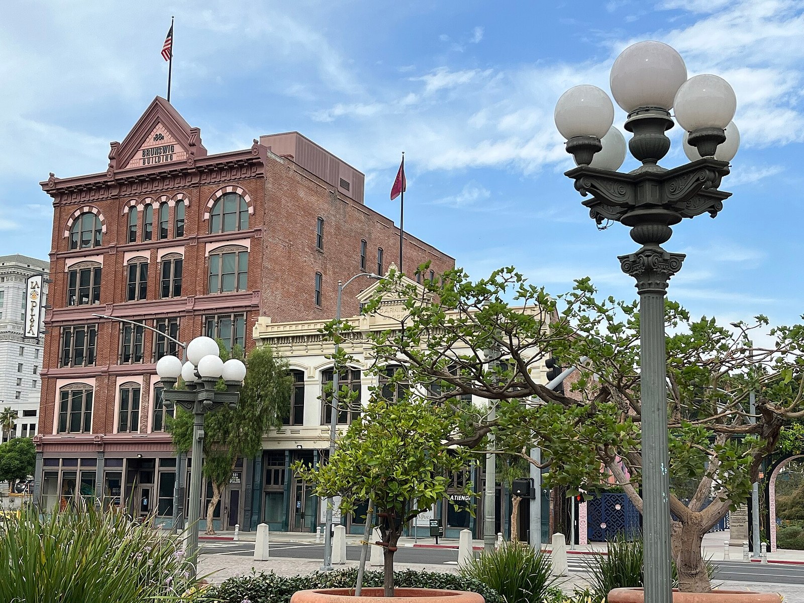 A green cast-iron lamp post topped with five white glass globes stands in the foreground, with a second matching lamp post, a small tree and a brick building behind it under a partly cloudy sky.