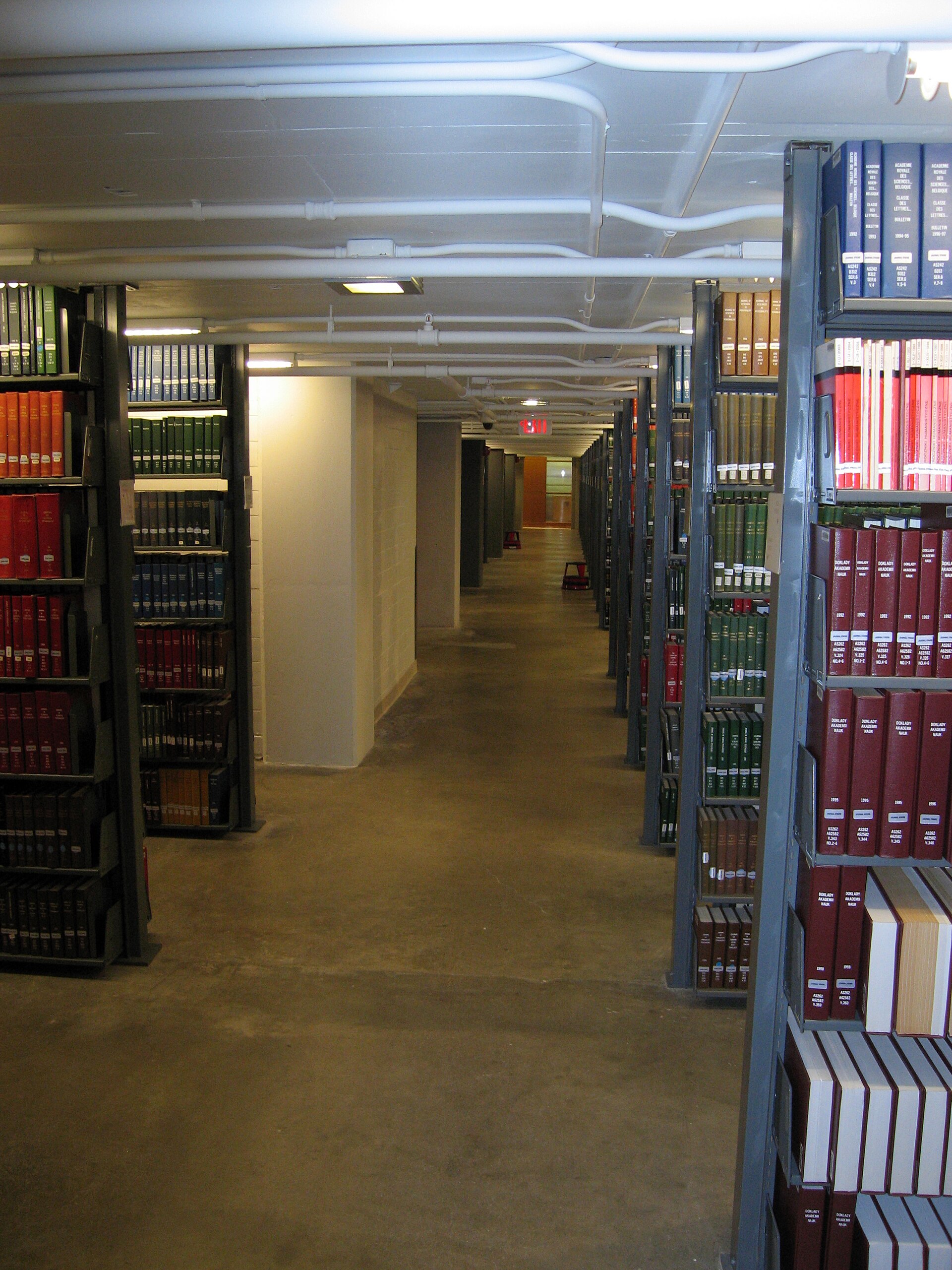 Rows of bound books and journals shelved on tall metal library stacks, an aisle receding between them under exposed ceiling pipes.