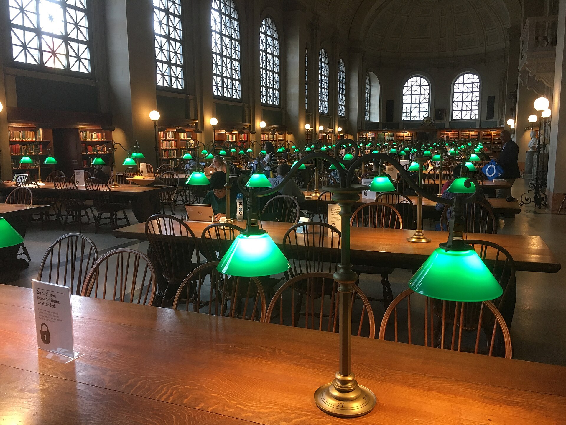 A grand library reading room with rows of long wooden tables lit by green-shaded brass lamps, tall arched windows and a coffered ceiling, a few people reading at the tables.