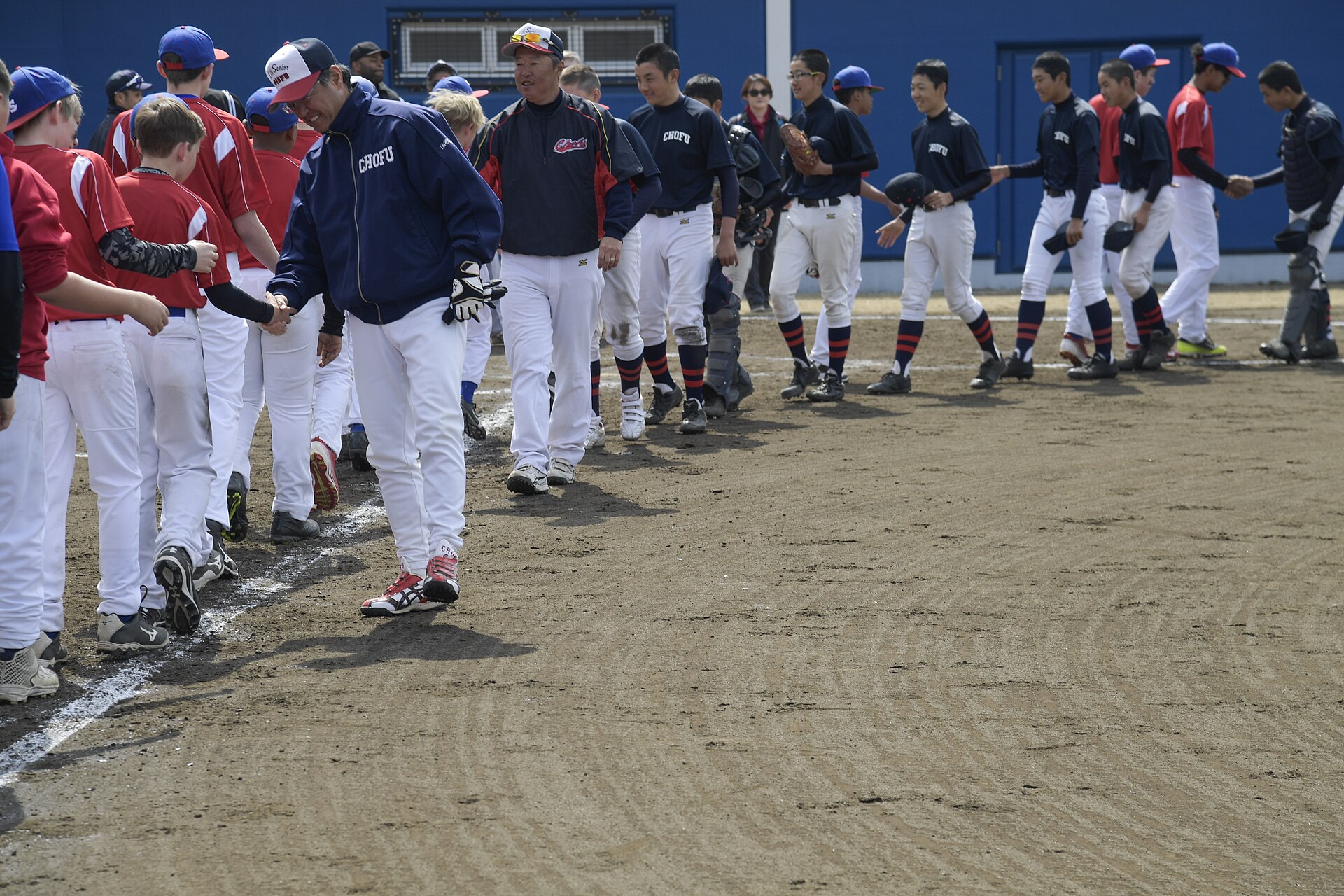 Two youth baseball teams and their coaches shake hands in a line across a dirt infield after a game.