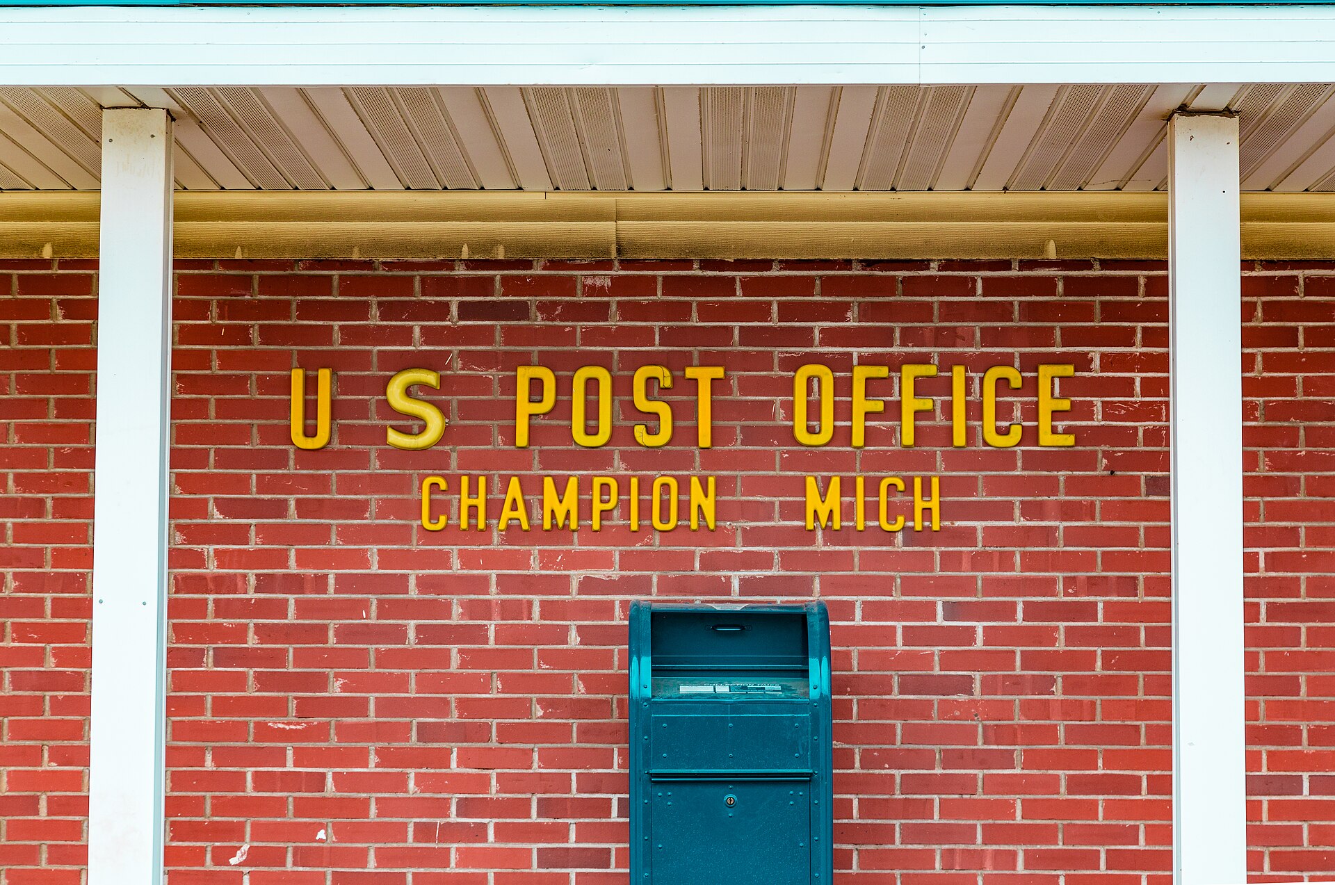A blue U.S. Postal Service mail collection box stands against a red brick wall beneath raised metal letters reading US Post Office.