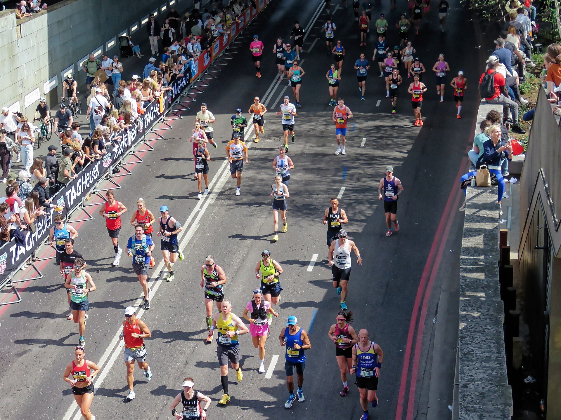Hundreds of numbered runners fill a wide, sunlit road passing through a cutting, spectators lining barriers on the left and sitting along a ledge on the right.