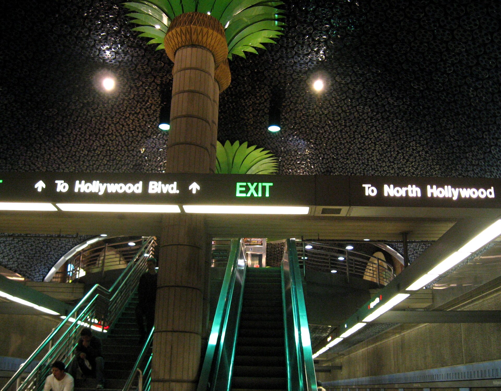 Escalators and stairs rise beneath decorative palm-tree columns and a film-reel ceiling inside the Hollywood/Vine subway station, below signs reading To Hollywood Blvd. and To North Hollywood.