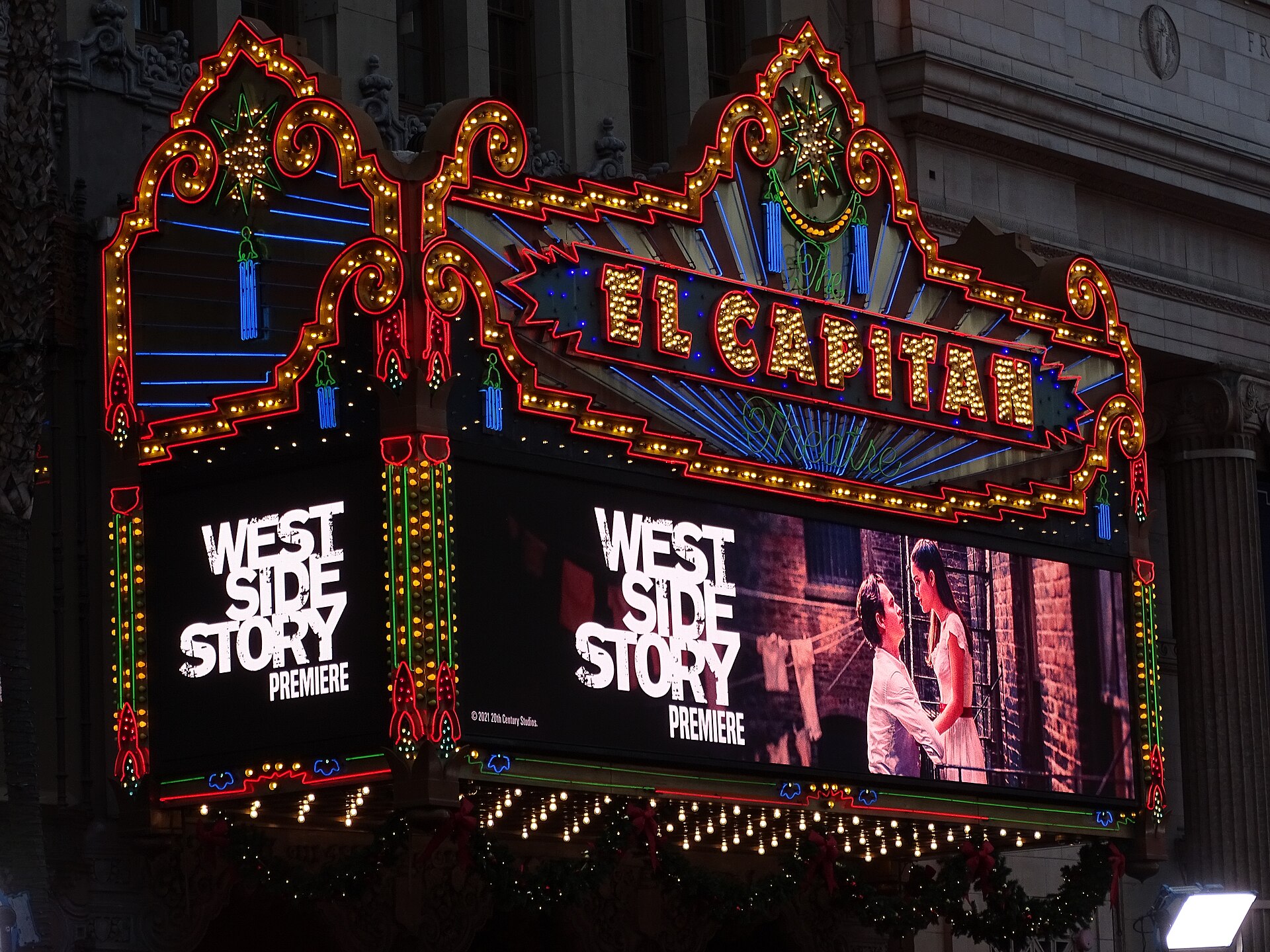 The El Capitan Theatre's ornate neon-and-bulb marquee glows red, gold and blue at night above two lit panels advertising a film premiere.