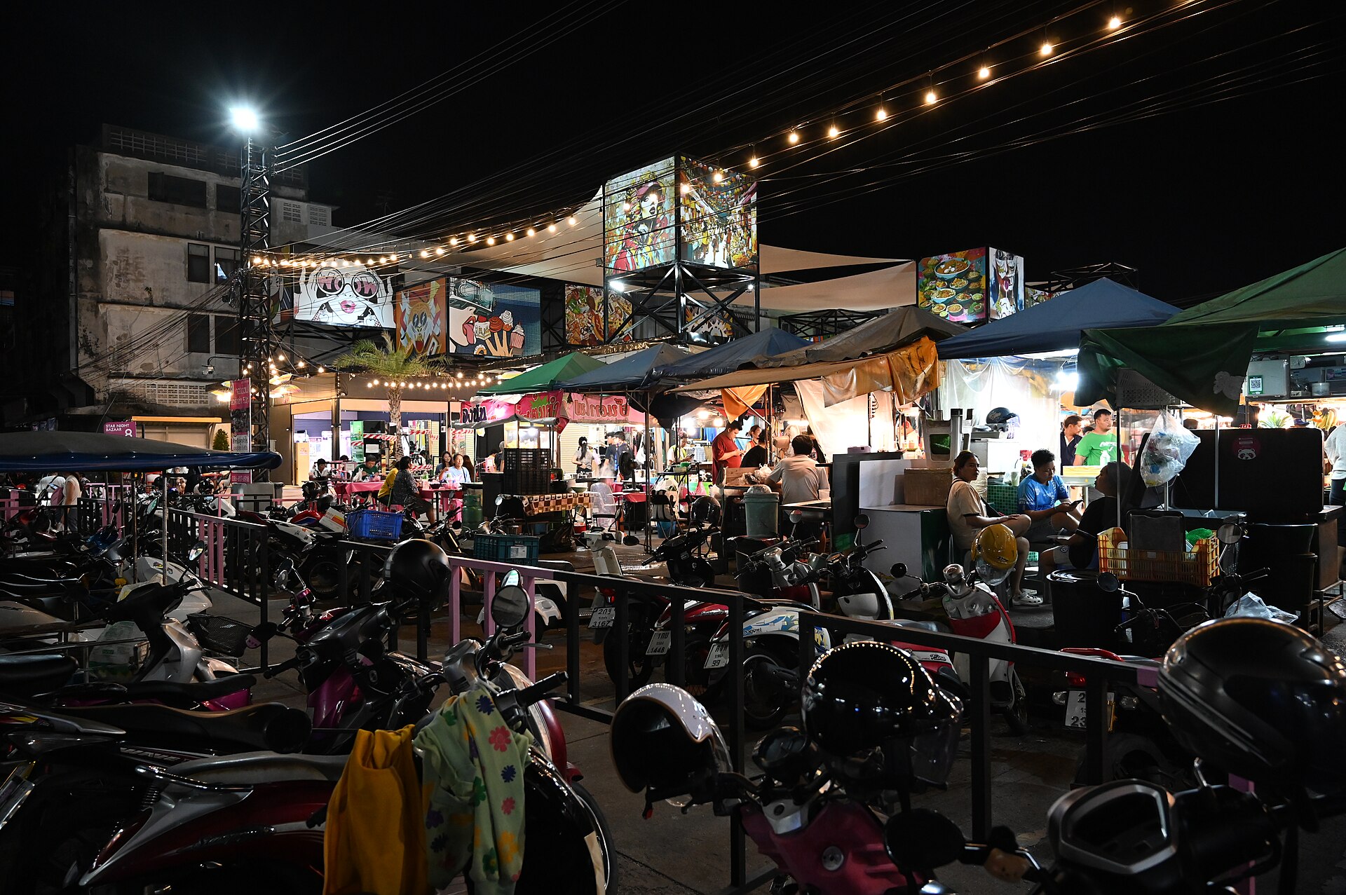 Rows of parked motorscooters stand behind a low railing in the foreground, with food stalls under canopies and strings of overhead lights at an outdoor market at night.