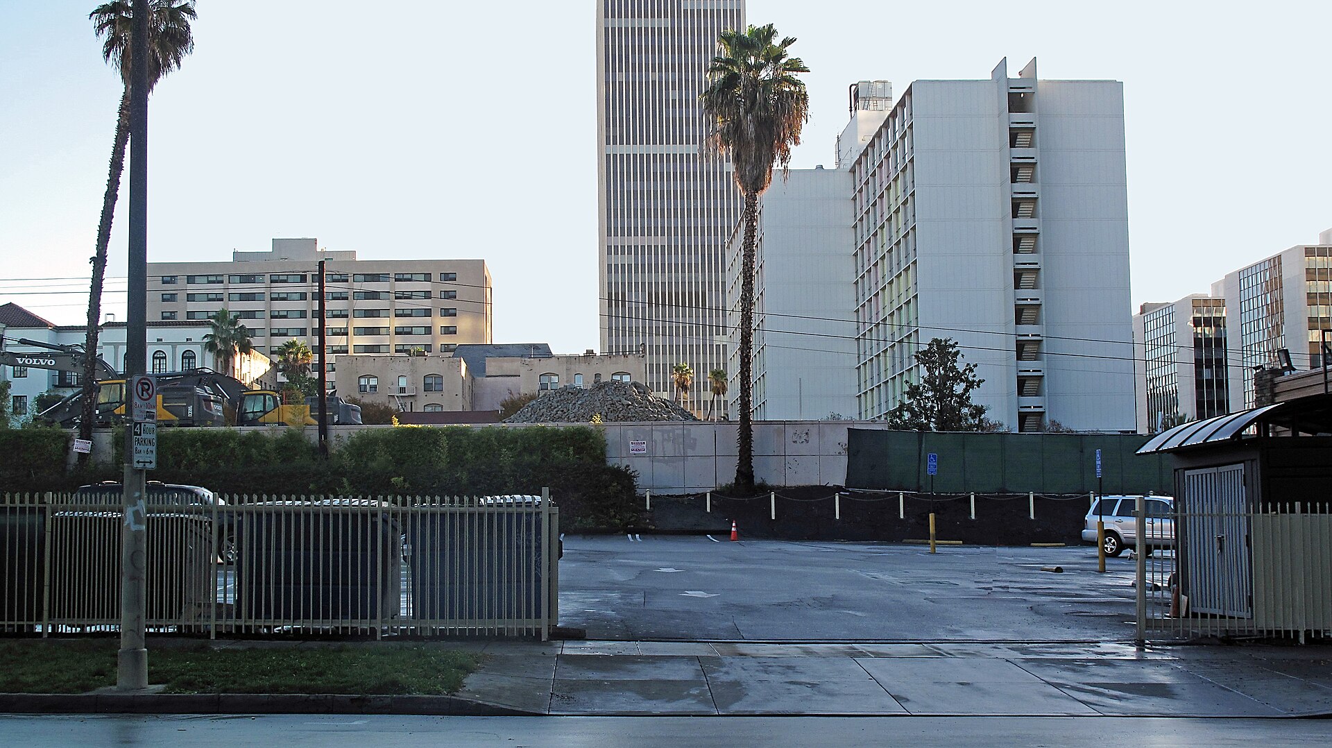 A mostly empty asphalt surface parking lot marked with white stalls sits behind a low wall, a commercial building and utility poles rising behind it.