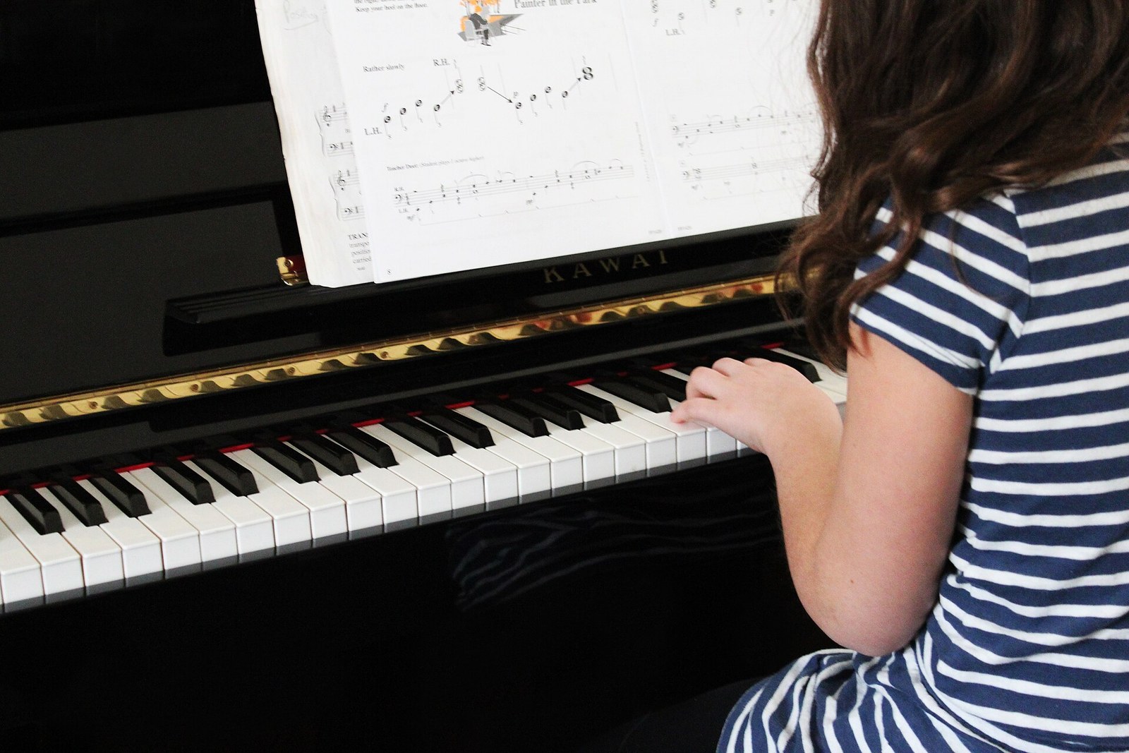 A girl with long brown hair and a striped top, seen from behind, plays a black upright piano with a beginner's music book open on the stand.