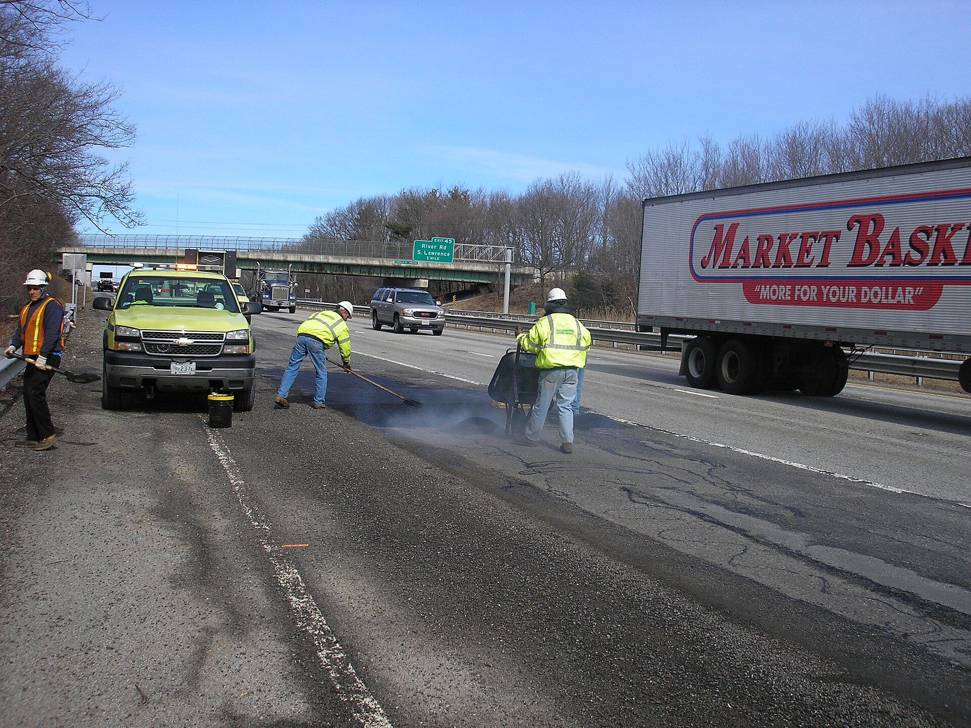 A road crew in hi-vis vests fills and rakes hot asphalt patch on a highway lane as traffic passes on the far side.