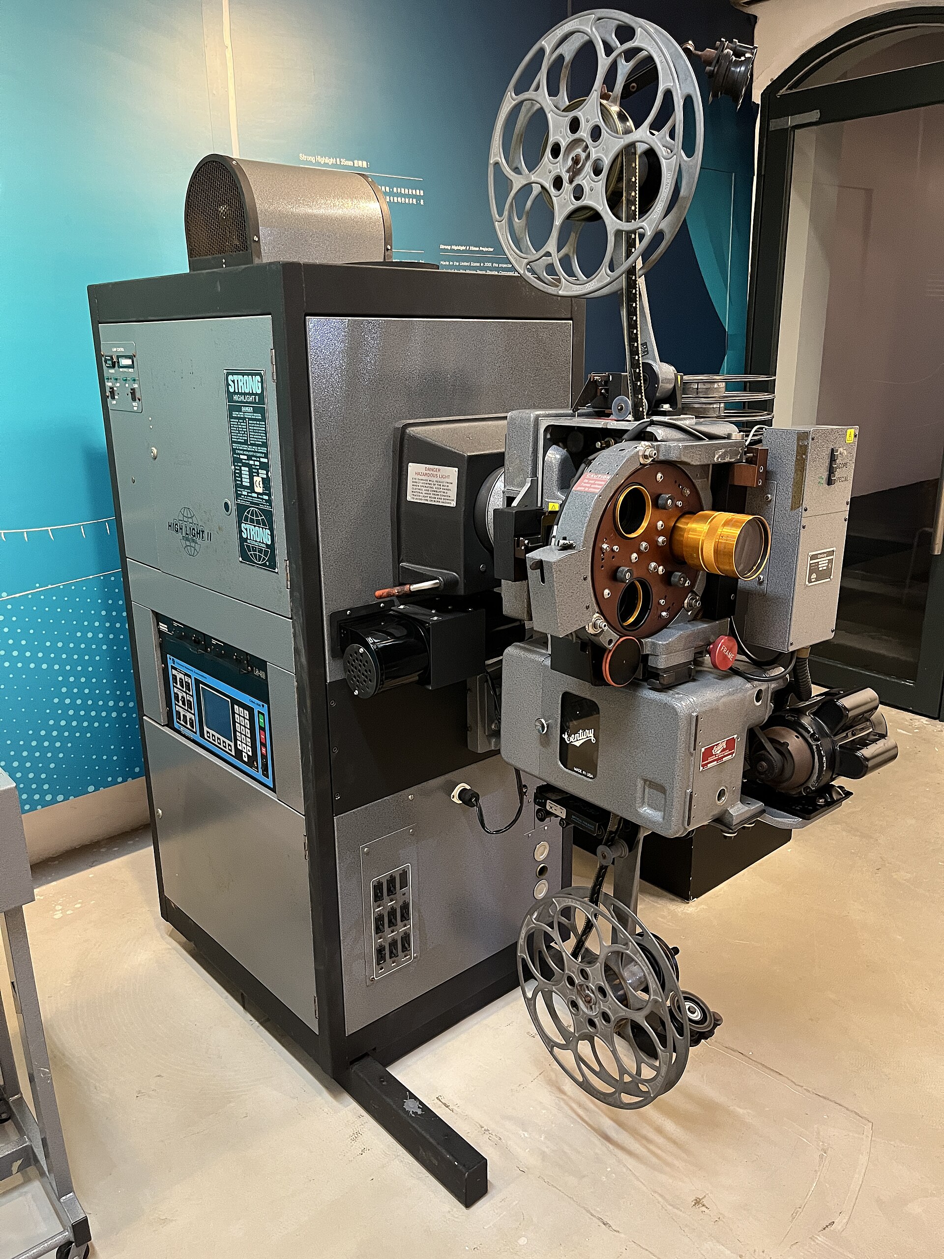 A large 35-millimeter film projector with tall metal film reels above and below, a lamphouse labeled "Strong Highlight II" and a lens, set against a teal exhibit wall.