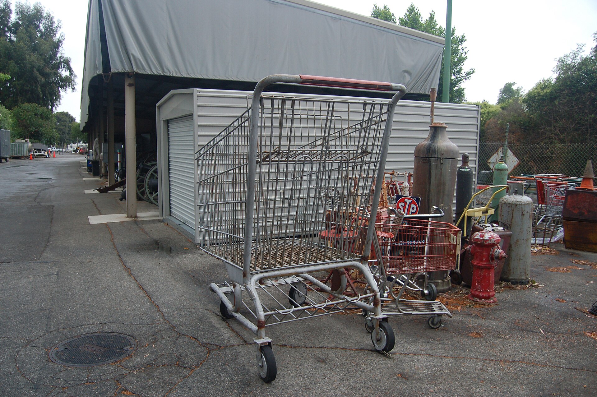 A large metal shopping cart and other salvaged objects — old gas cylinders, a red fire hydrant and an "STP" sign — sit on asphalt outside a covered storage structure.