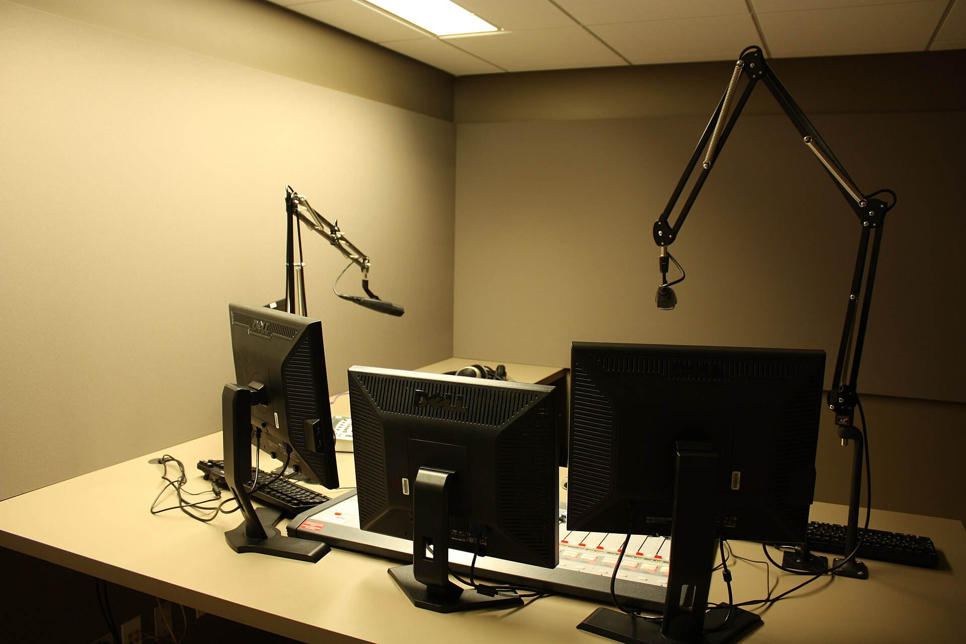 A radio broadcast studio desk with an on-air mixing console, three computer monitors and two large microphones on articulated boom arms in a plain room.