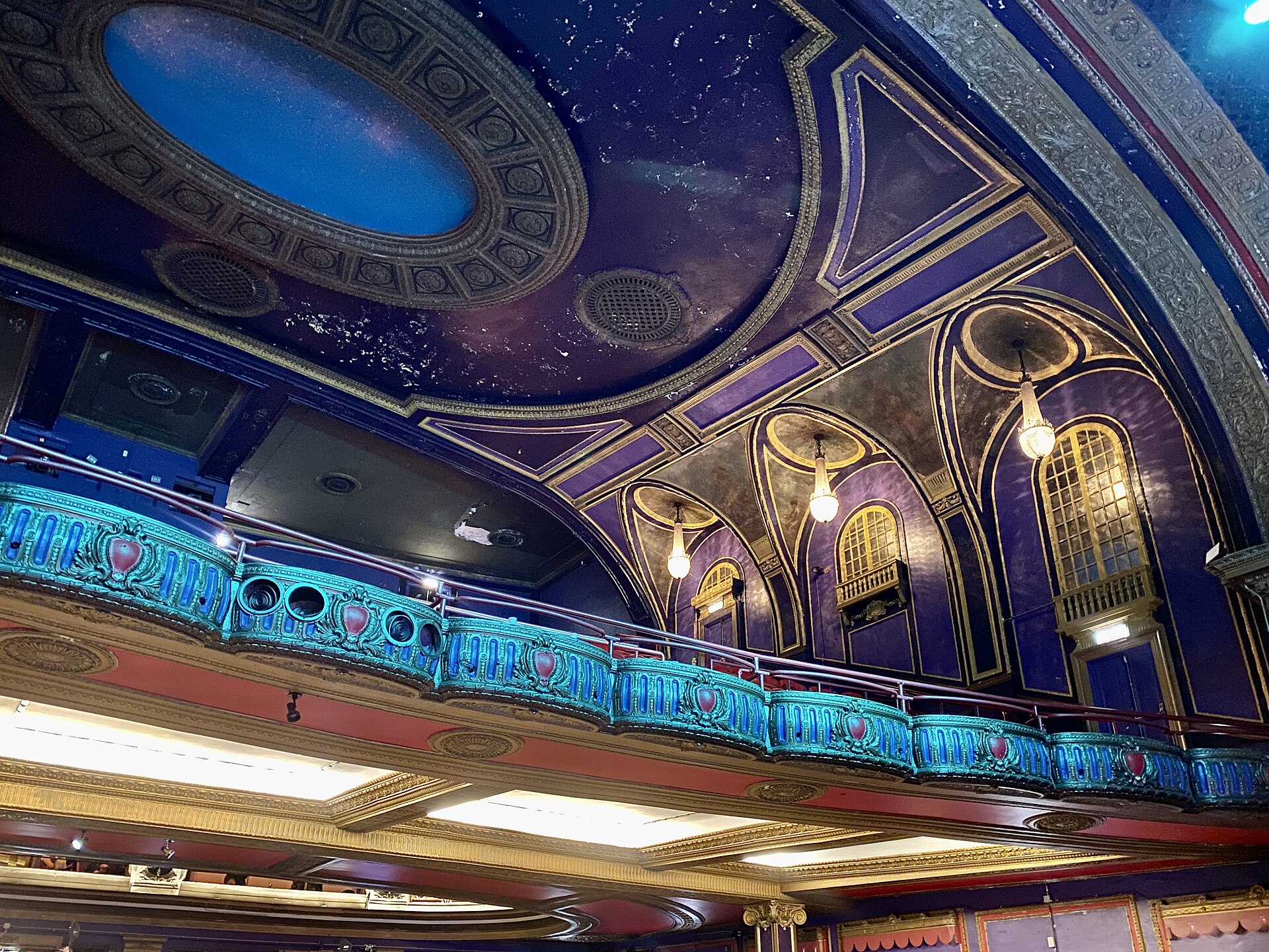The ornate balcony and painted dome ceiling of a historic movie palace, with turquoise decorative panels, gilded arched windows and hanging light fixtures.