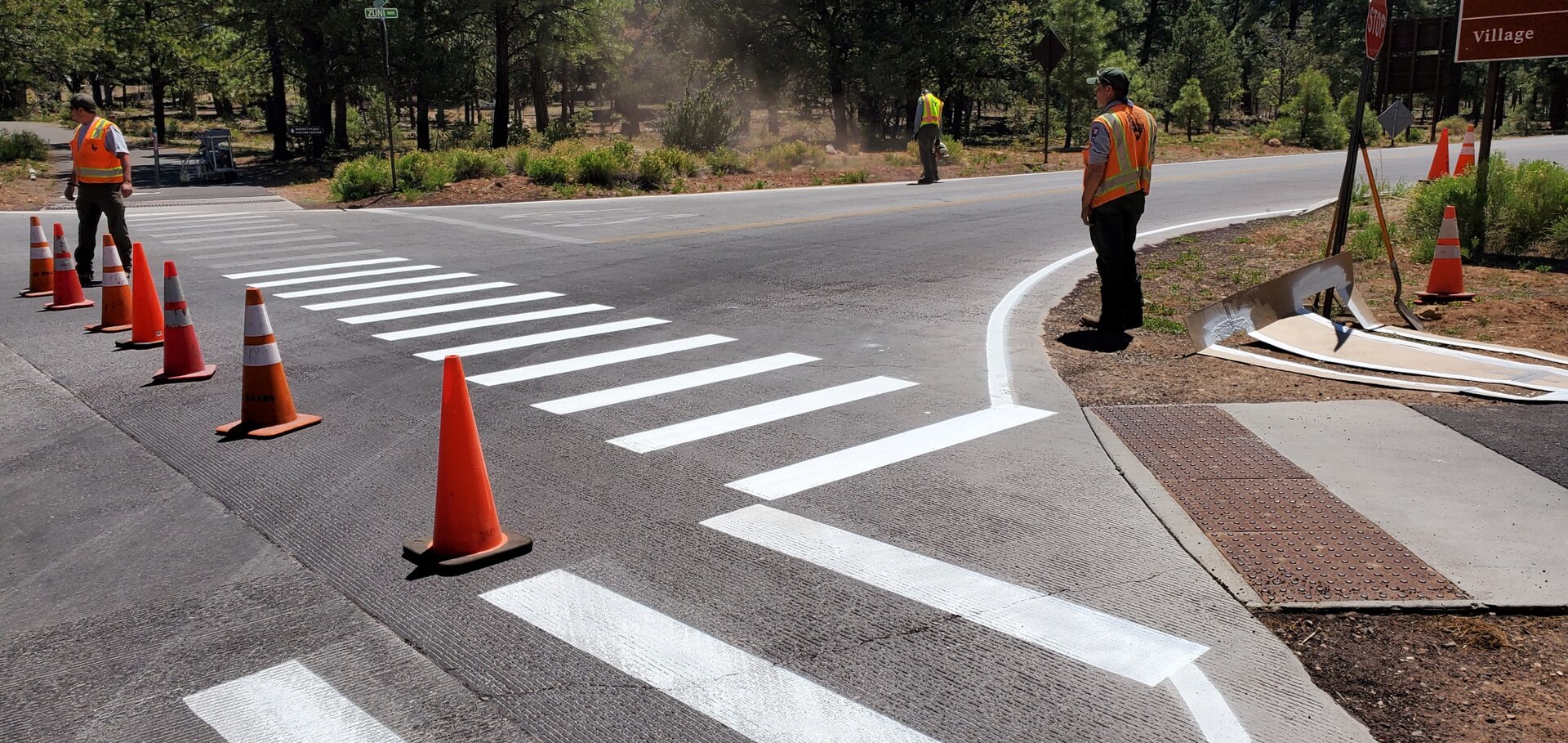Three road-crew workers in orange safety vests stand around a freshly painted white crosswalk and curb line at a tree-lined intersection, with orange traffic cones and a stencil board lying beside the wet paint.